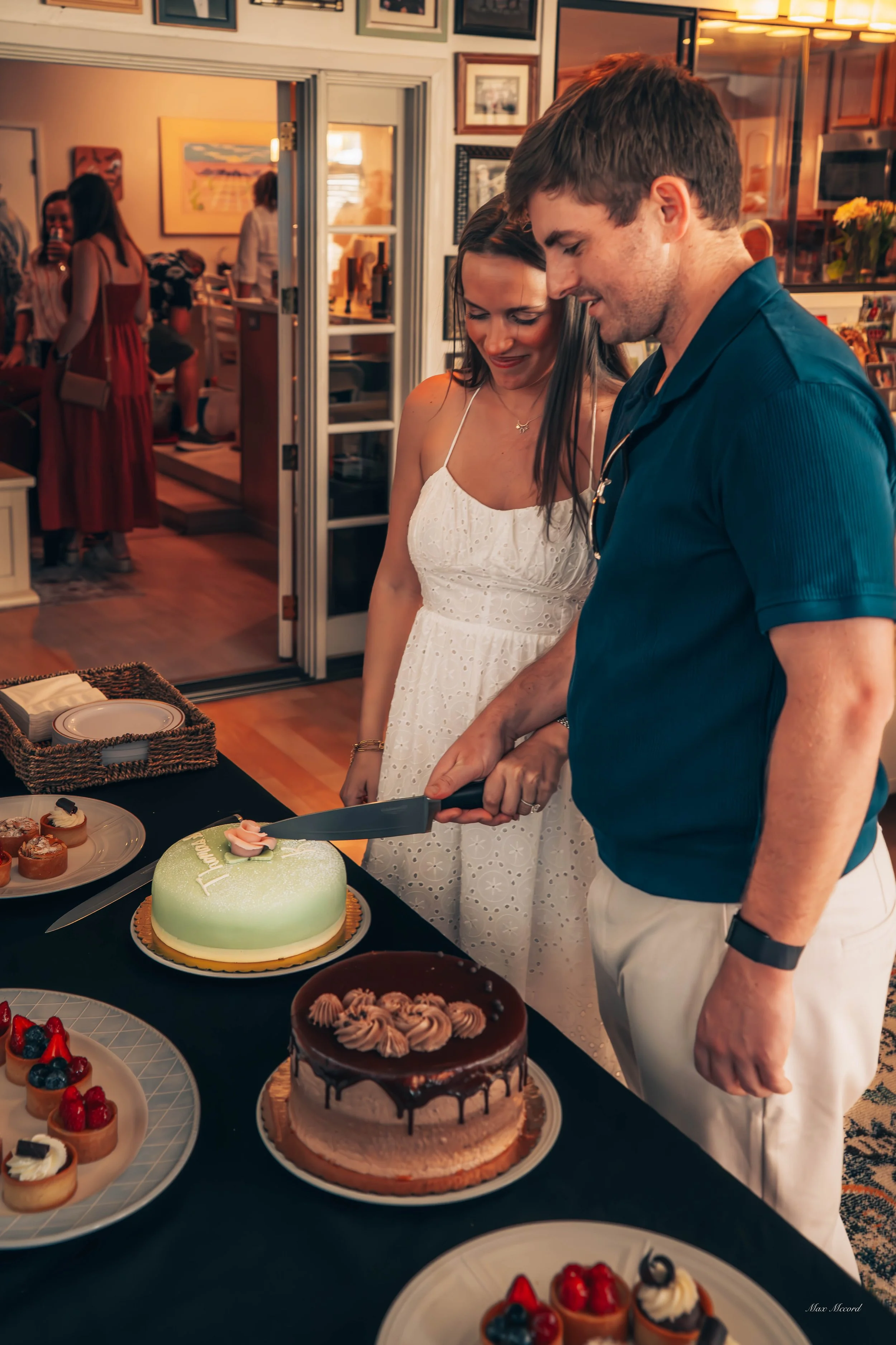 A man and woman cutting a birthday cake at a celebration. The lady wears a white dress and the man a navy blue shirt. There are various desserts on the table, and people in the background.