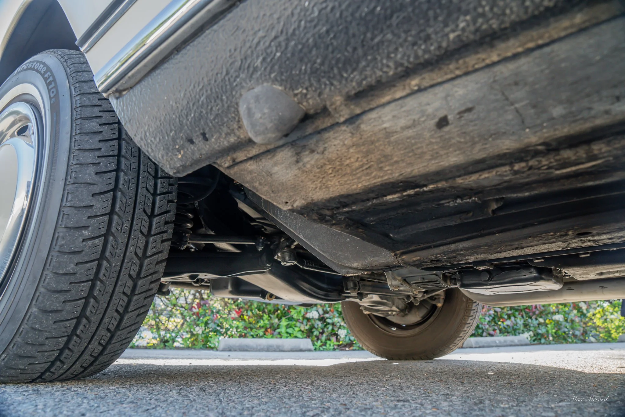 Underside view of a vehicle showing tires, suspension, and undercarriage with pavement and bushes in background.