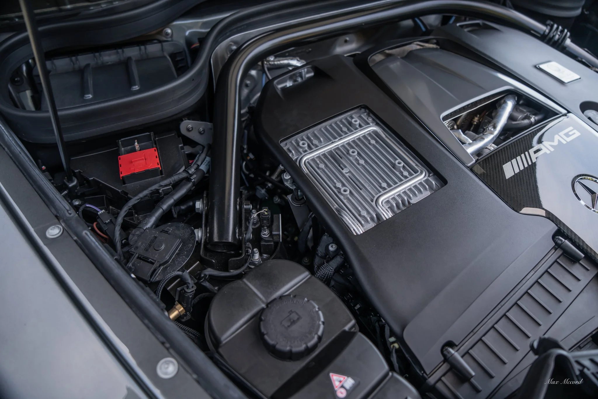 Close-up of a car engine bay with Mercedes-Benz AMG branding, showing various engine components, hoses, and metal parts.