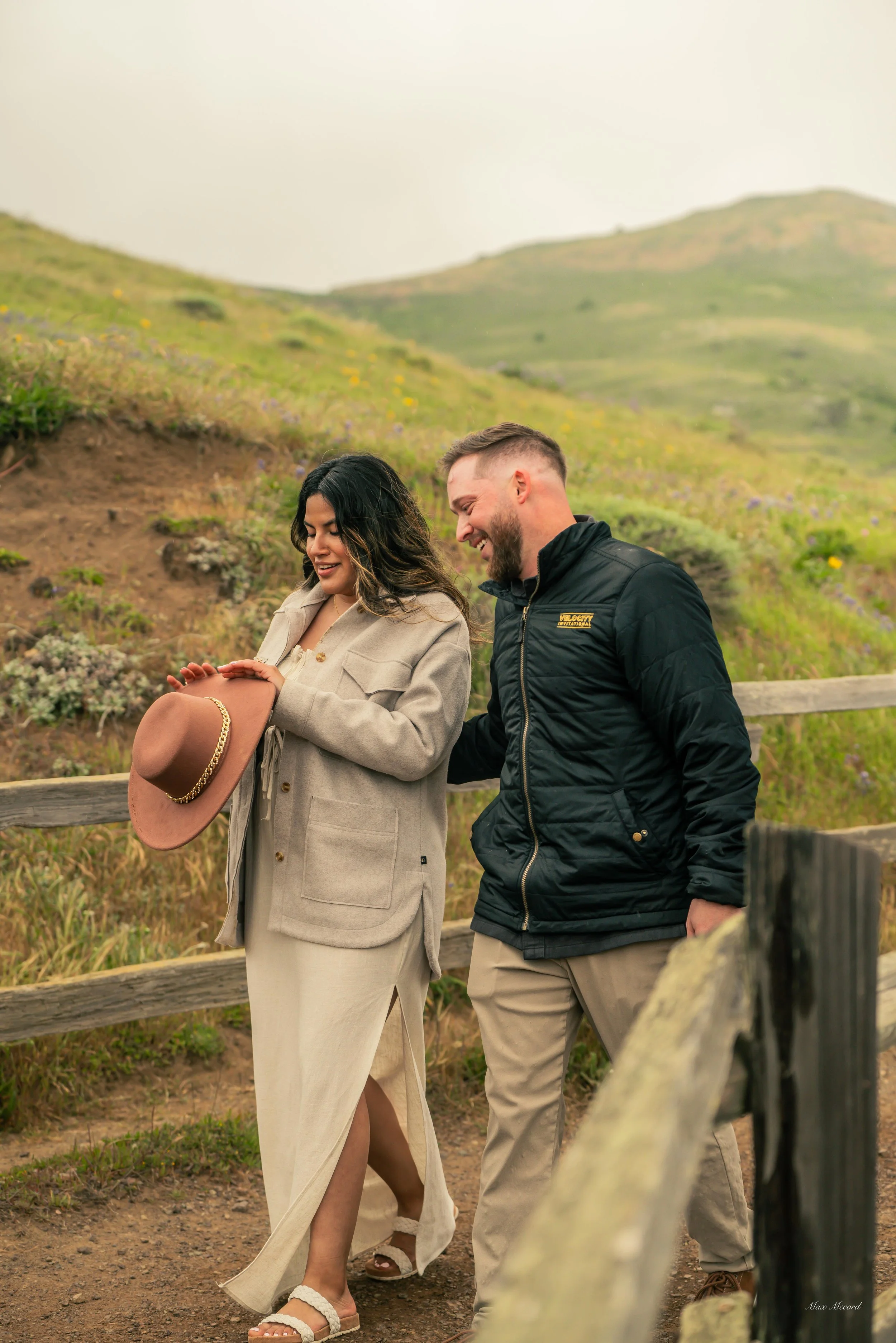 A man and woman smile and walk together on a trail with a grassy hillside in the background. The woman holds a hat while the man gently touches her arm.