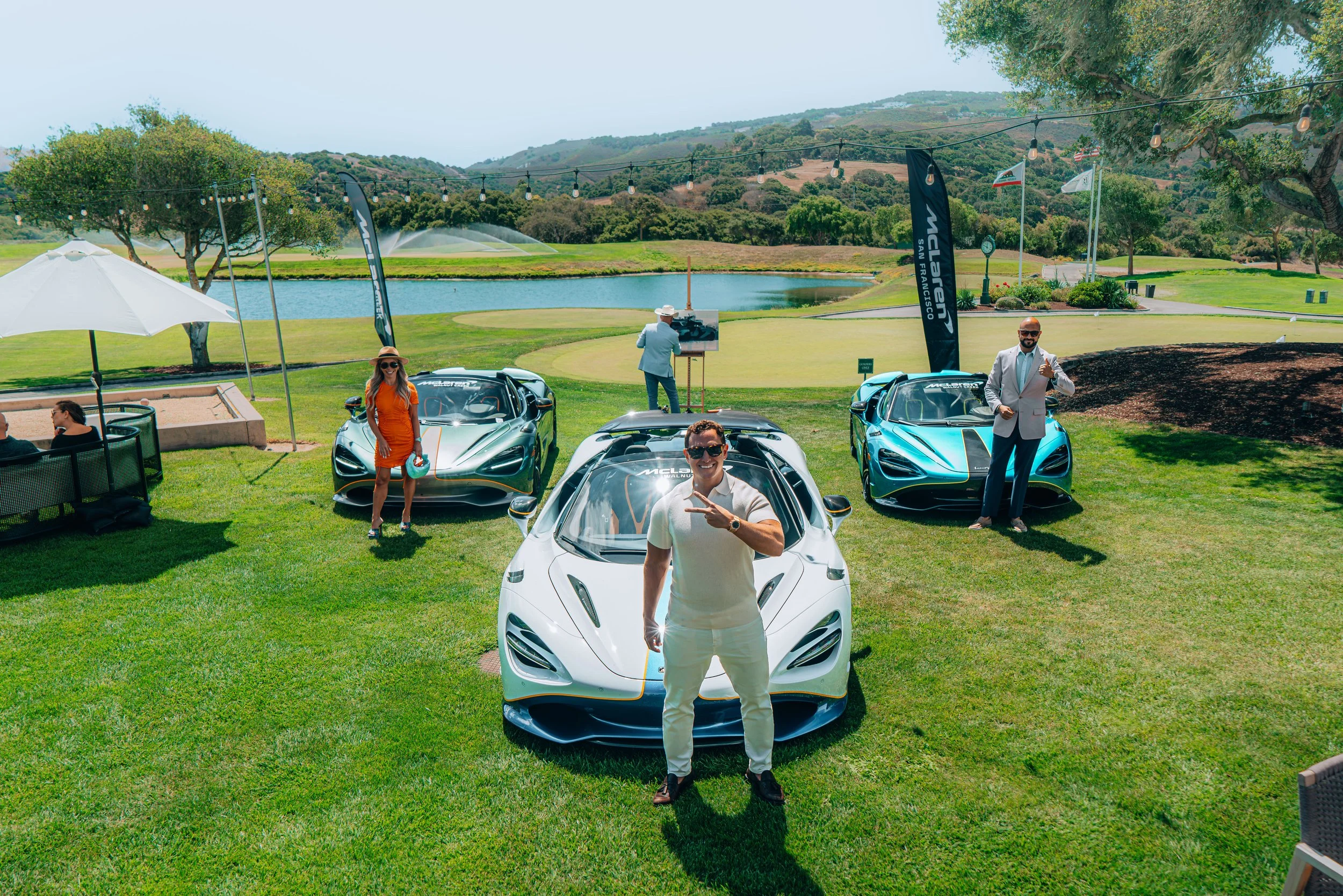 Three people stand in front of high-performance sports cars on a lush golf course with water features and rolling hills in the background. One man in a white shirt in the foreground is smiling and gesturing with a peace sign. A woman in an orange dre