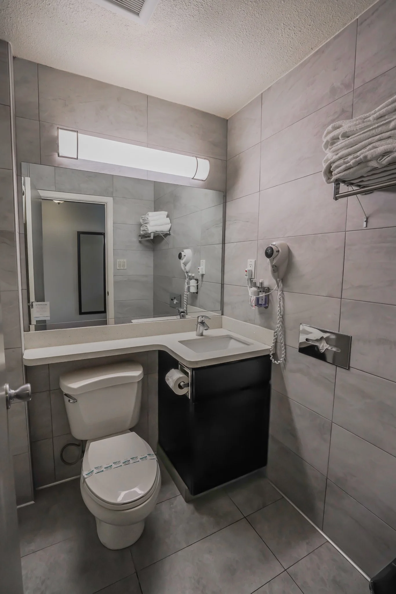 A small modern bathroom with a toilet, a sink, a large mirror, and wall-mounted hairdryers. Towel racks with folded towels are above the sink. The walls and floor are tiled in gray.