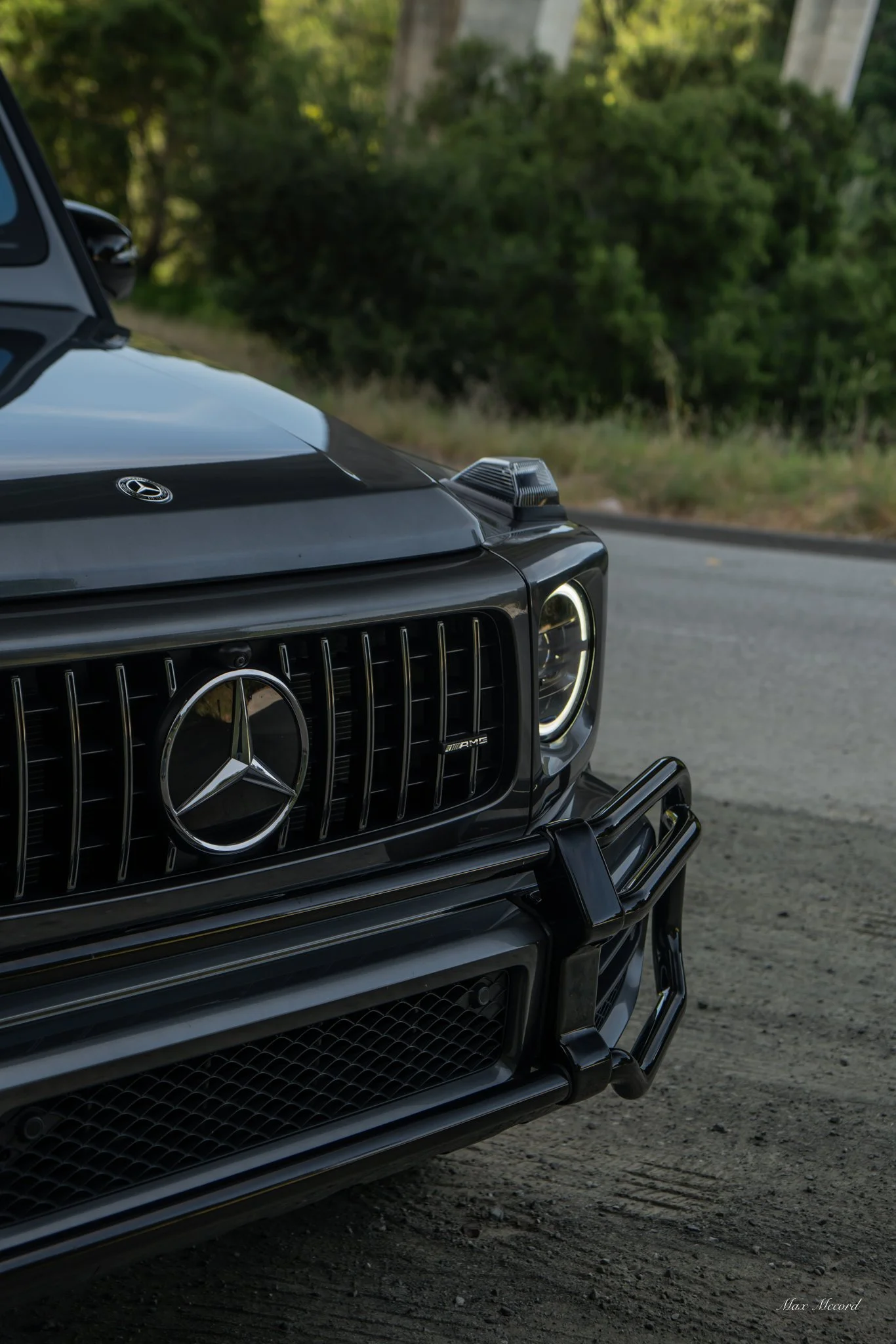 Close-up of the front of a black Mercedes-Benz G-Class SUV with Mercedes emblem and AMG badge, parked on a dirt roadside with greenery in the background.
