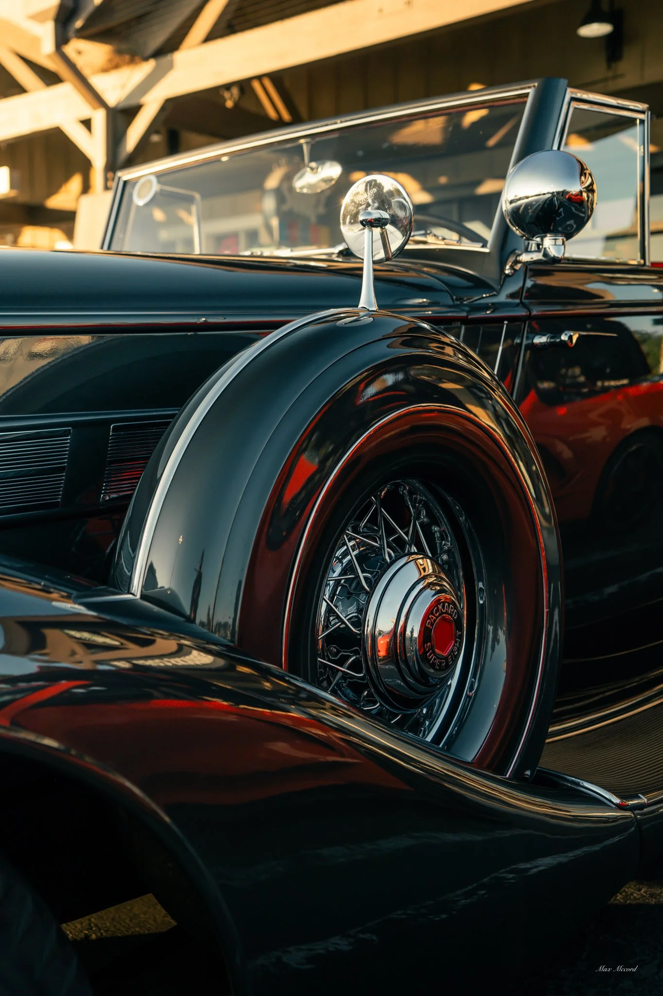 Close-up of a vintage black convertible car with chrome details, including wire-spoked wheels and side mirrors, parked under a wooden structure during sunset.