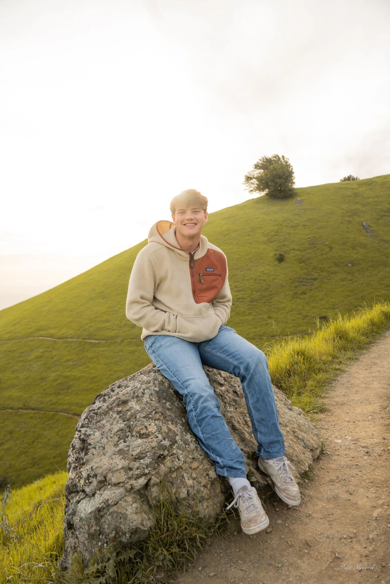 A young man sitting on a large rock on a grassy hillside, smiling at the camera during sunset, with a green hill and a single tree in the background.