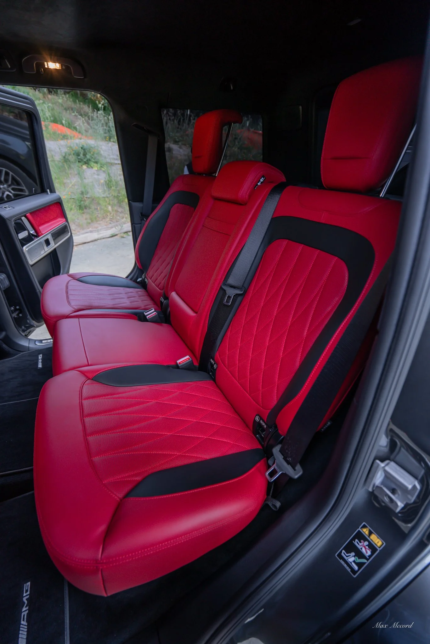 Interior of a vehicle showing row of three red and black leather seats with black seat belts.