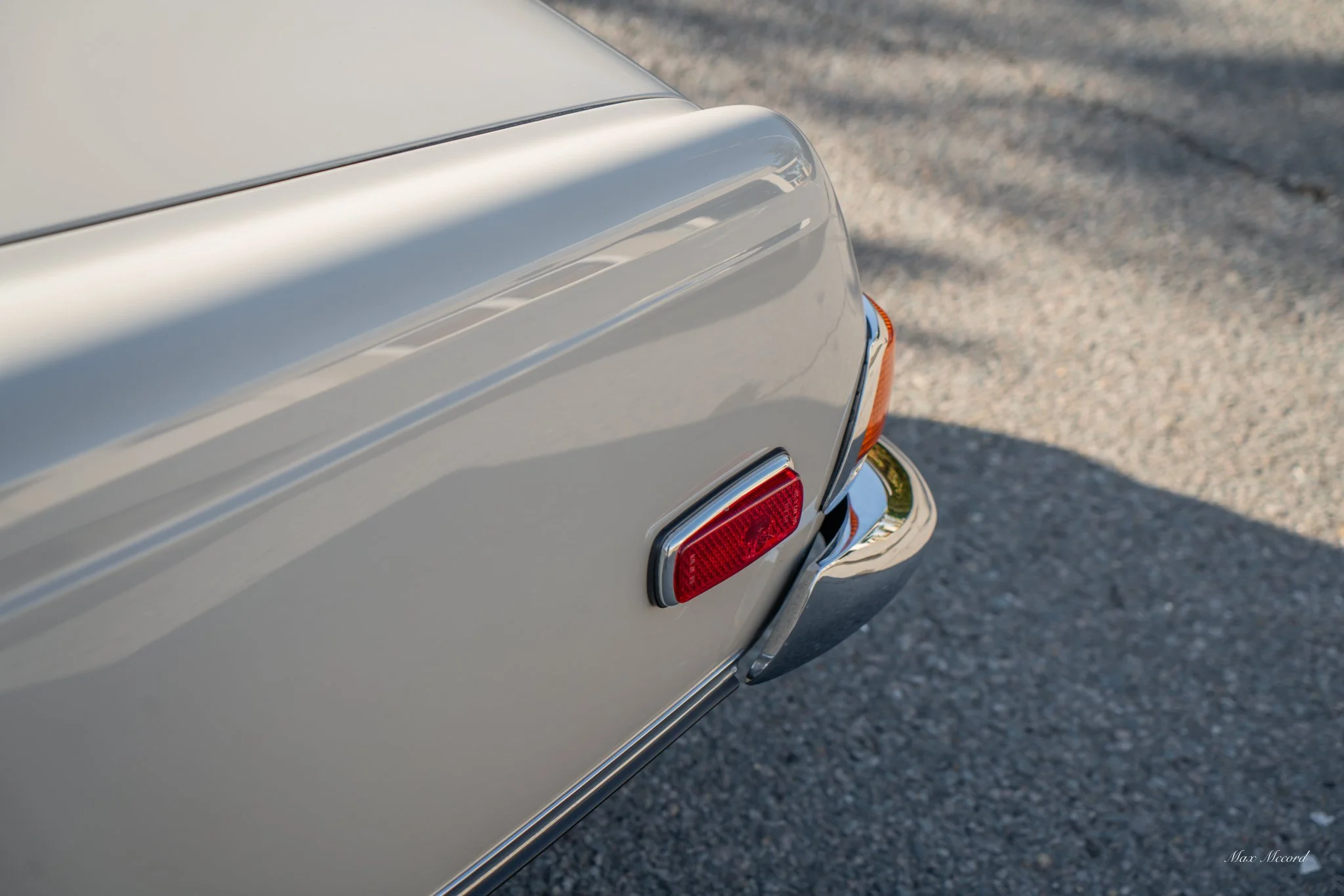 Close-up of the rear end of a vintage silver car with a red rear light and chrome bumper on a gravel surface.