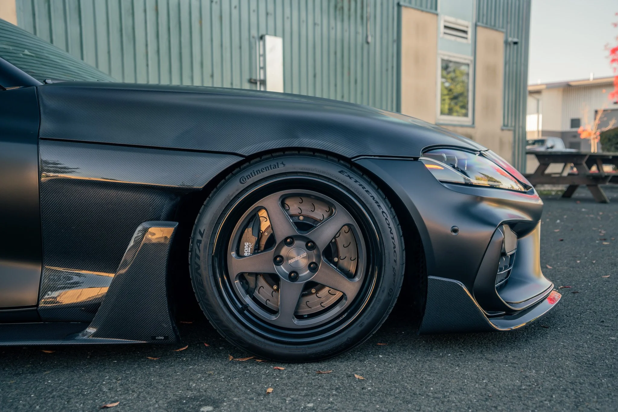 A black sports car with carbon fiber accents, featuring a racing wheel with Continental tires, parked on asphalt with a green building and outdoor picnic table in the background.