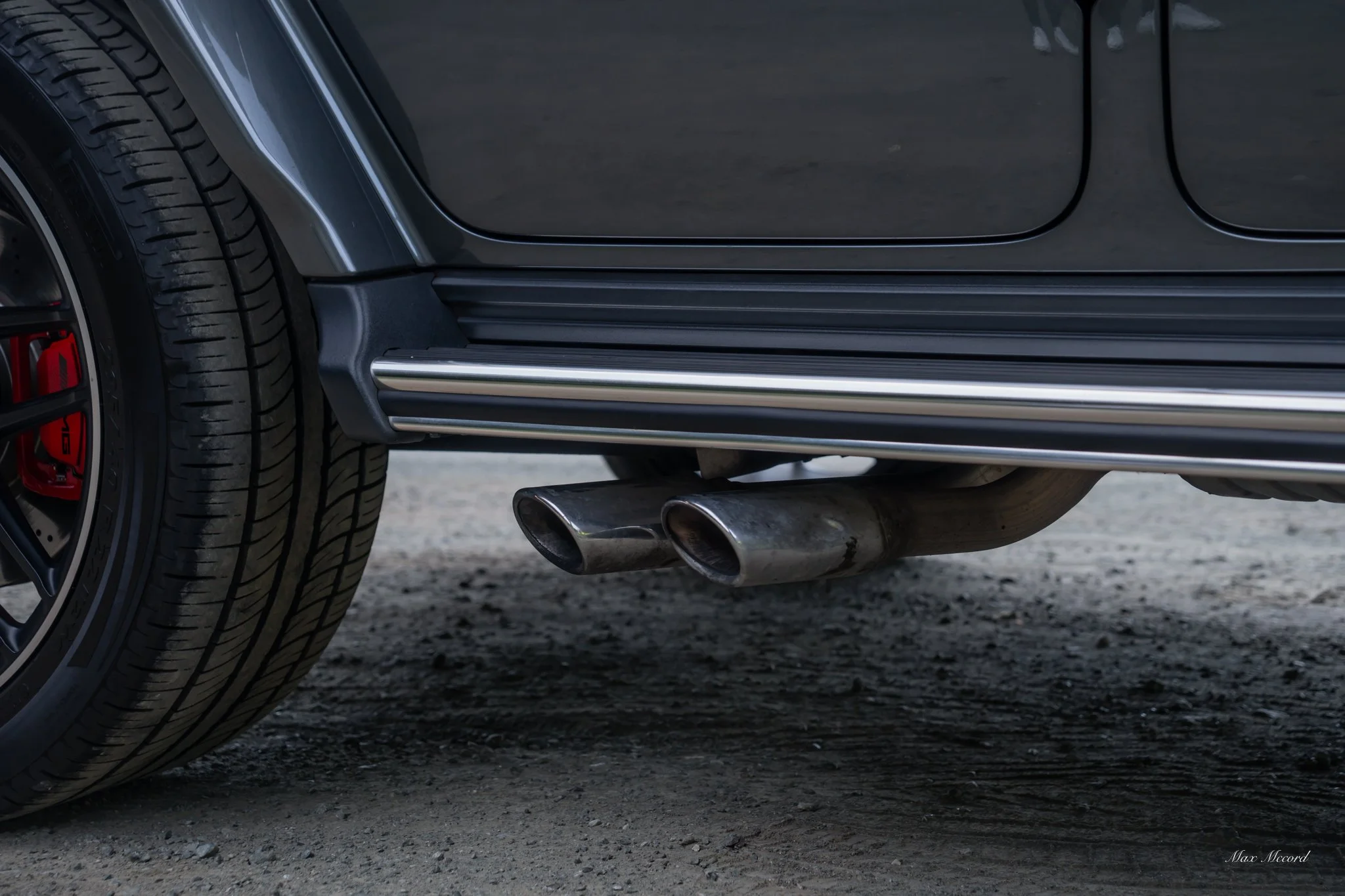 Close-up of a car's rear wheel and dual exhaust pipes on a gravel surface.
