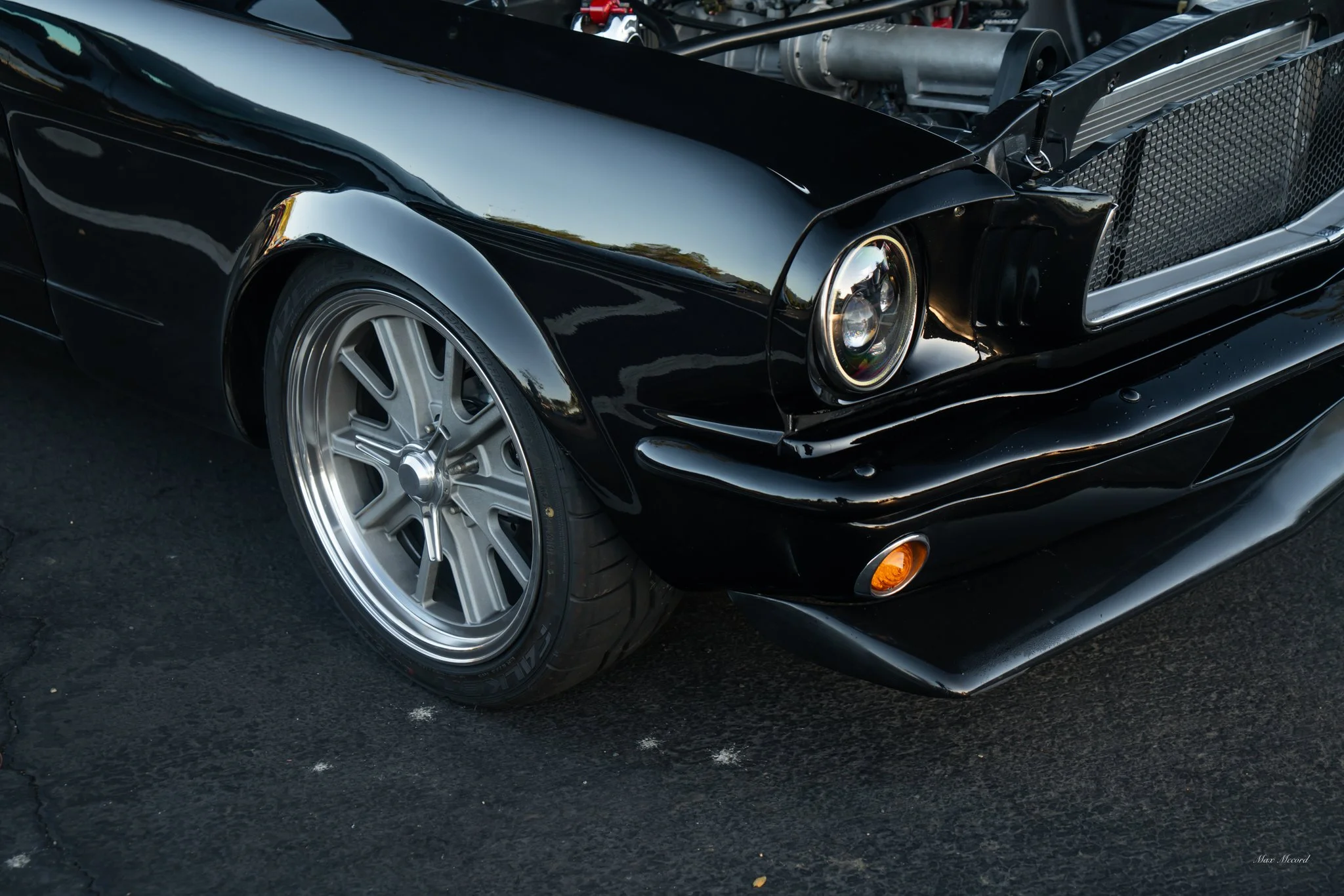 Close-up of the front left side of a black classic car with a polished silver wheel, single round headlight, and an orange turn signal on the bumper, parked on asphalt.