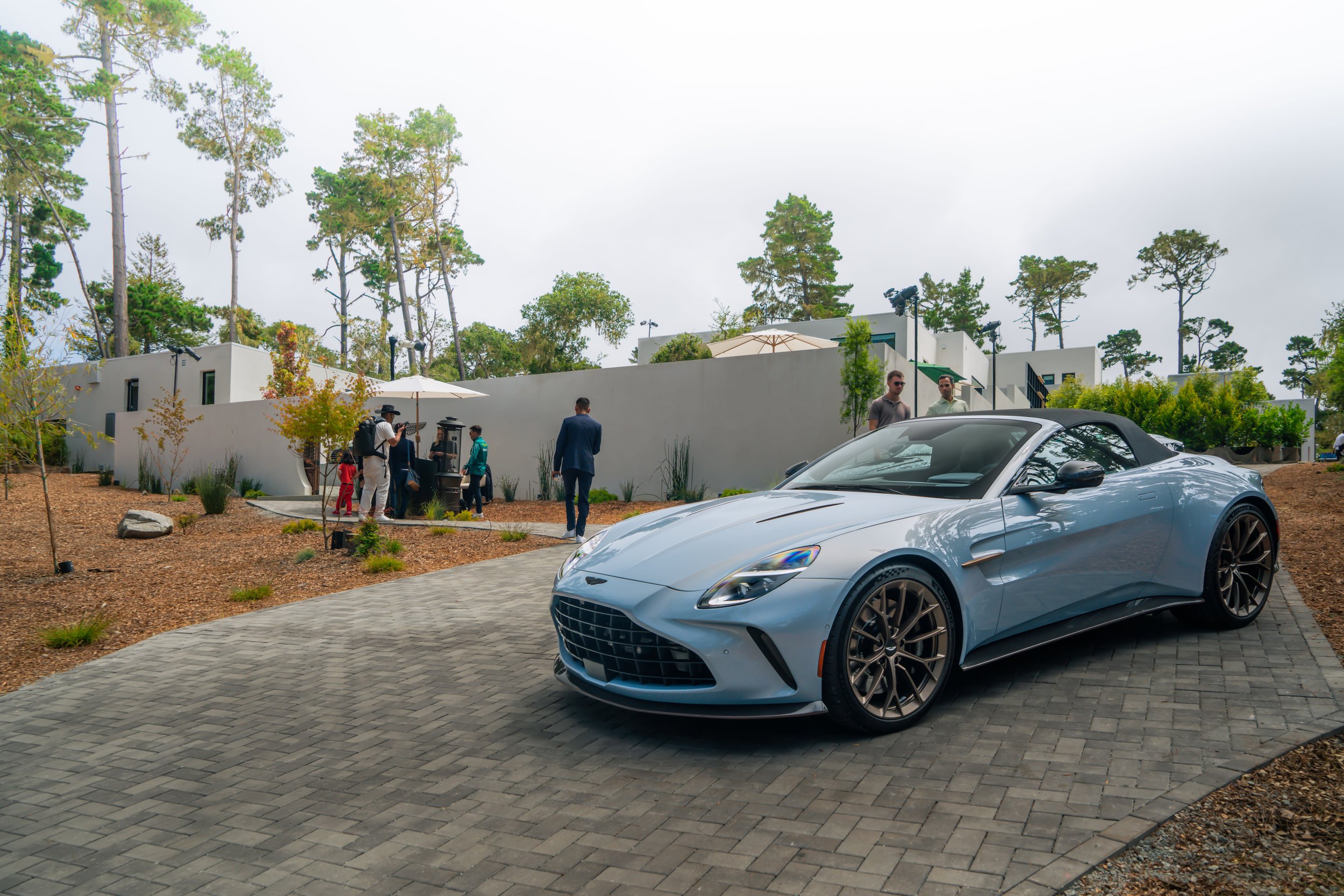 A silver sports convertible car parked on a cobblestone driveway outside a modern building with people gathered around, some talking and others walking, surrounded by trees and greenery.
