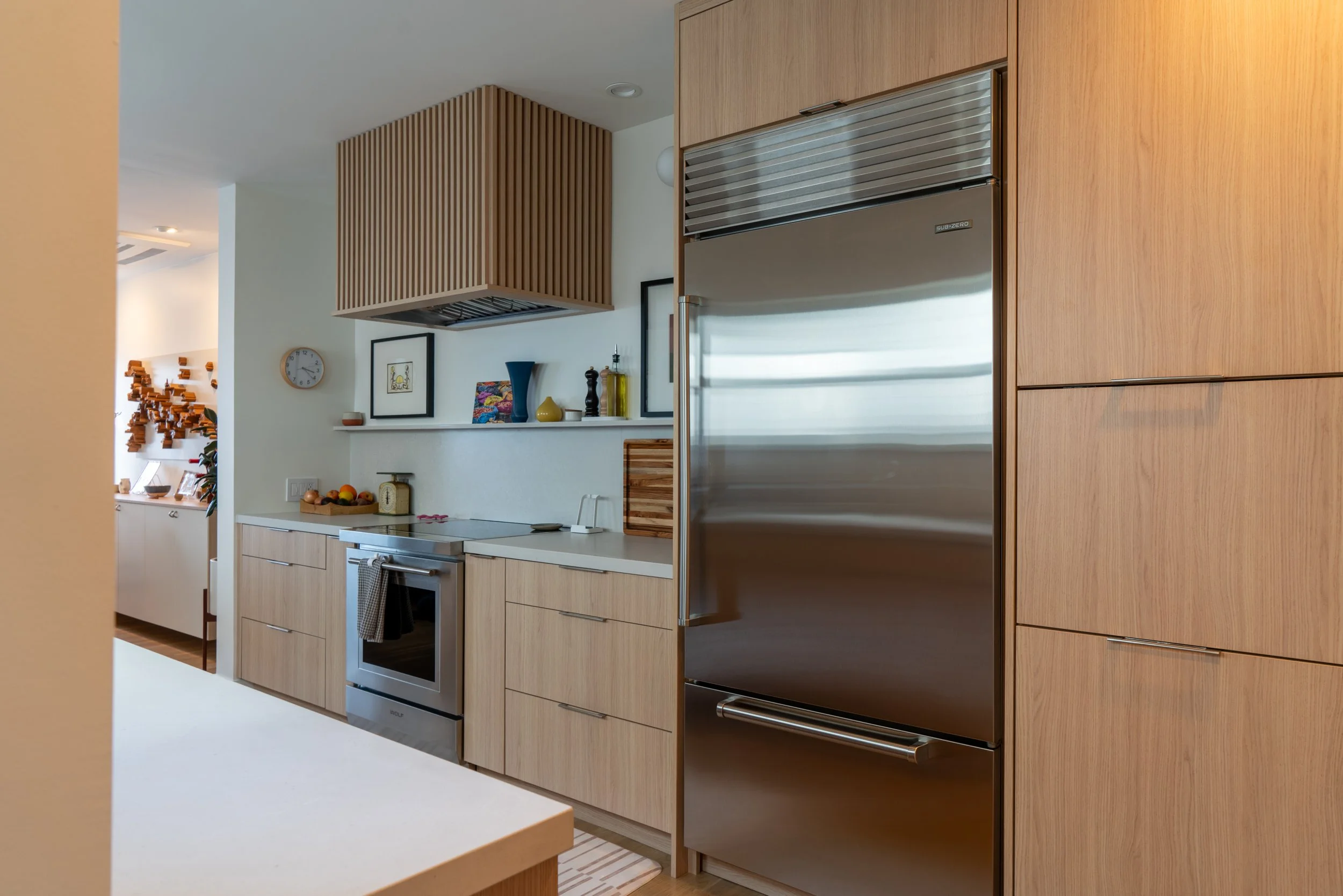 Modern kitchen with wooden cabinets, stainless steel fridge, white countertops, and decorative items on a shelf.