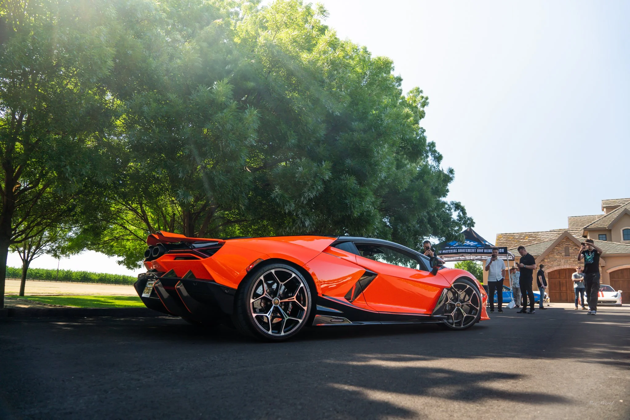 Orange sports car parked on the street with a group of people taking pictures and a house in the background.
