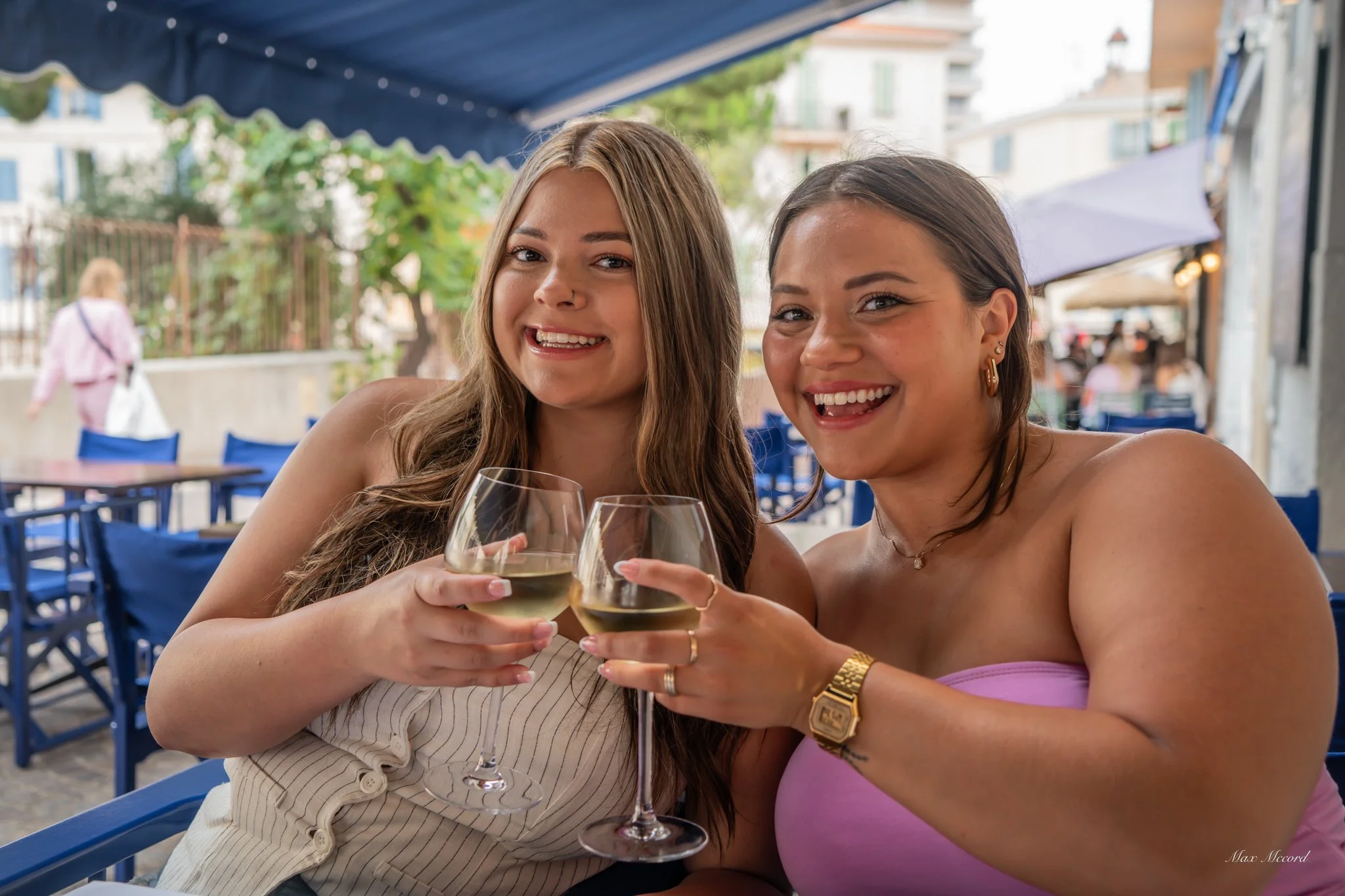 Two women smiling and clinking glasses of white wine at an outdoor cafe.