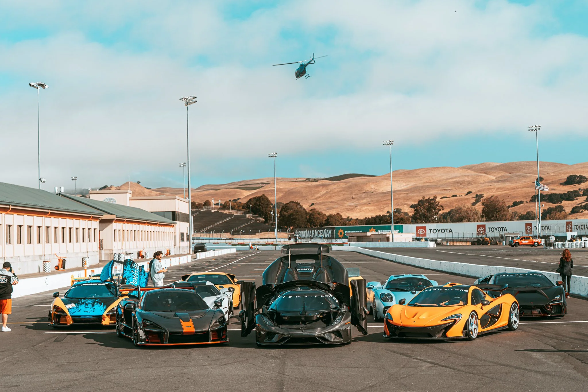 Several sports cars parked on a race track with a helicopter flying overhead. Hills in the background and people standing near the cars.