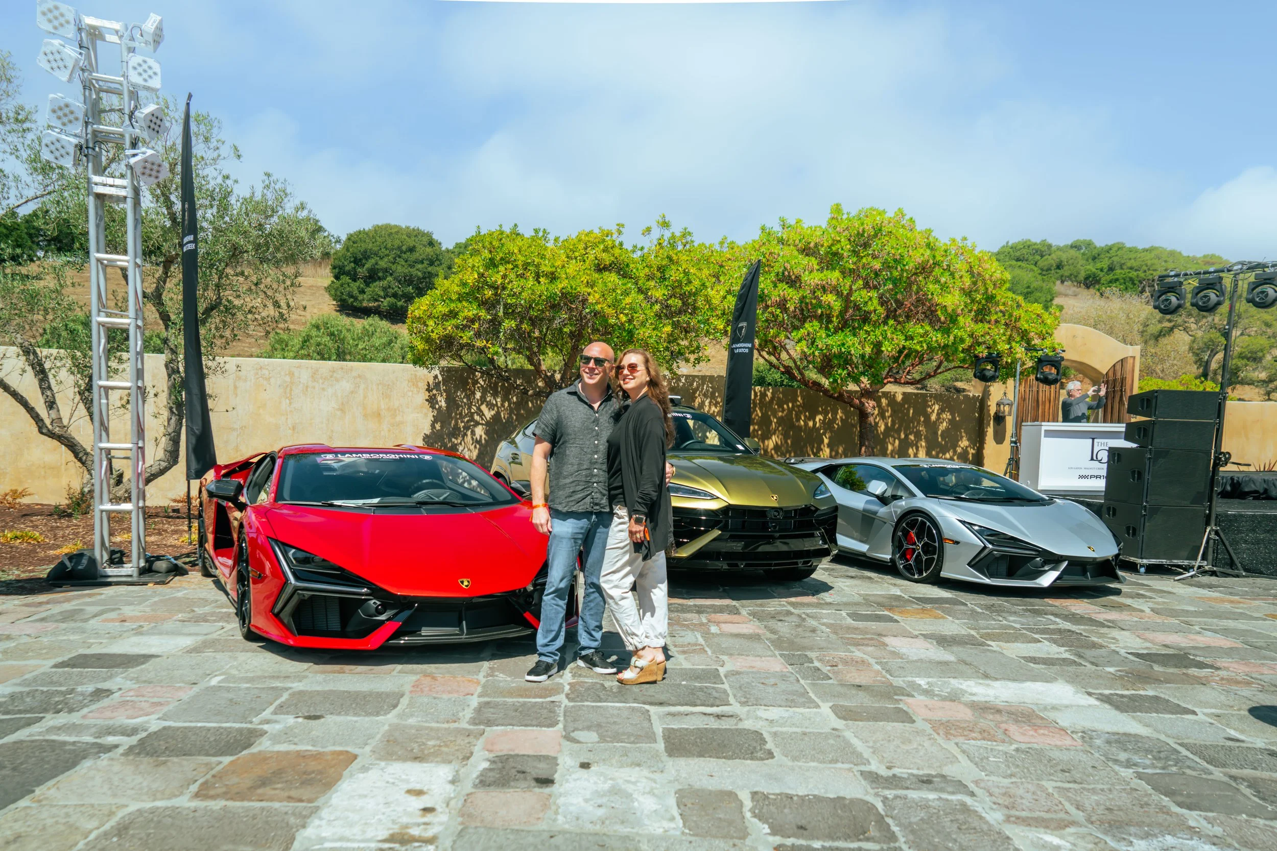 A man and woman stand together smiling in front of three luxury sports cars, including a red Lamborghini, a gold Lamborghini, and a silver Lamborghini, at an outdoor car event on a stone paved area with a backdrop of trees and a beige wall.