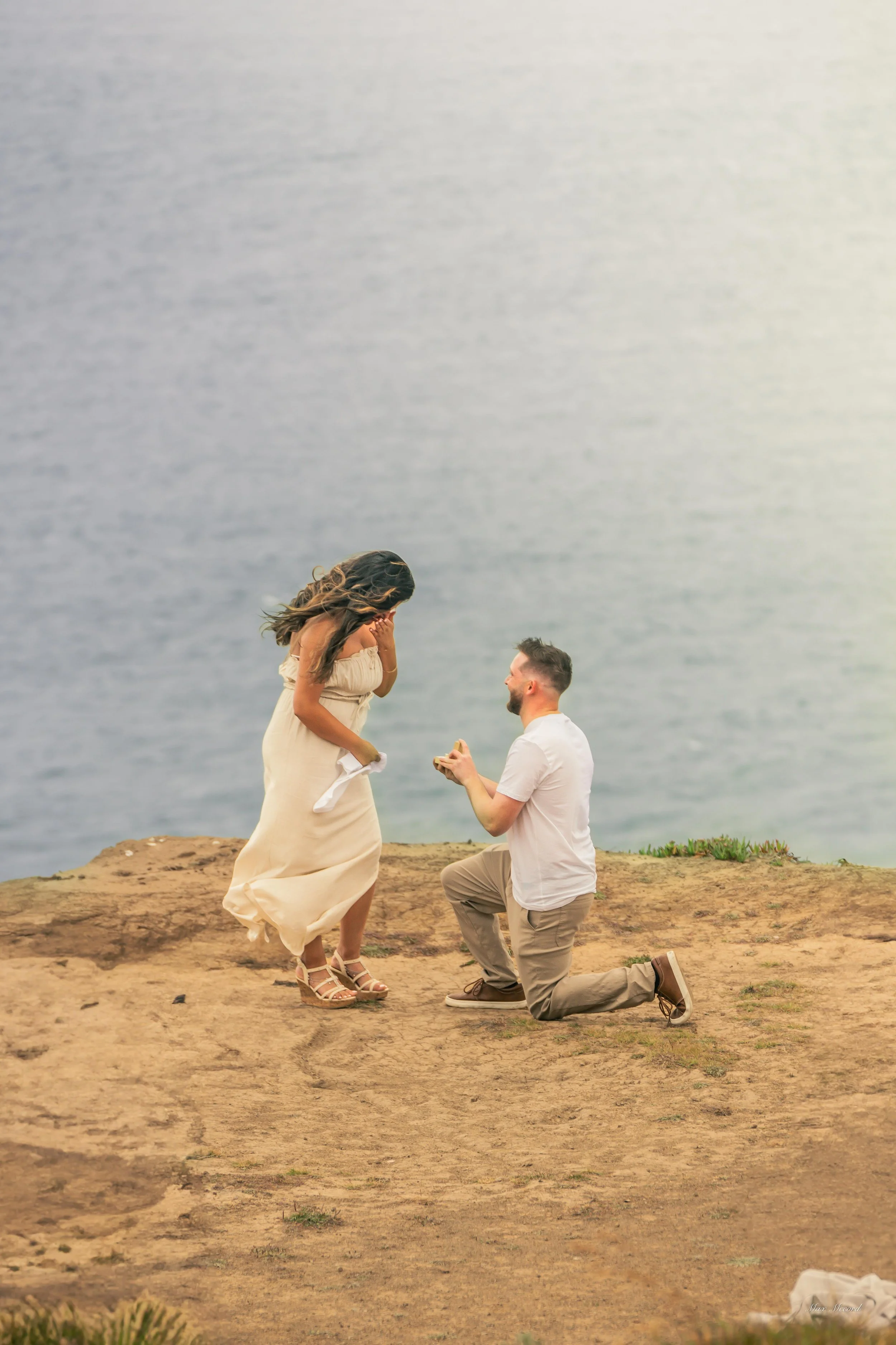 A man is kneeling on one knee proposing marriage to a woman standing in front of him on a cliff overlooking the water.