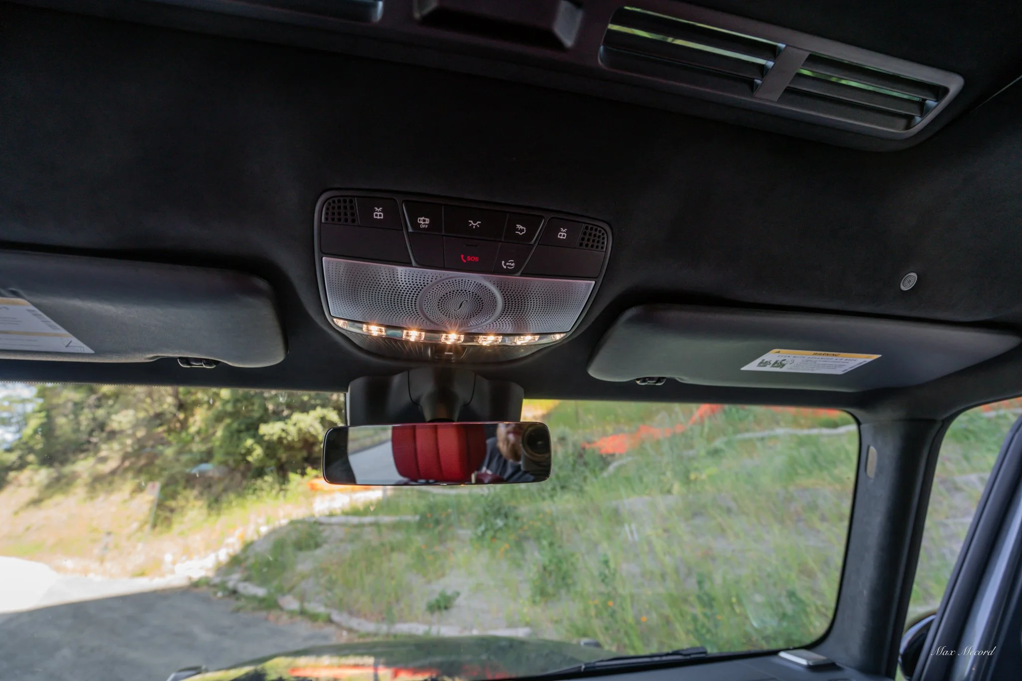 View of the interior roof of a vehicle showing overhead control panel, rearview mirror, and sun visors with a scenic outdoors visible through the windshield.