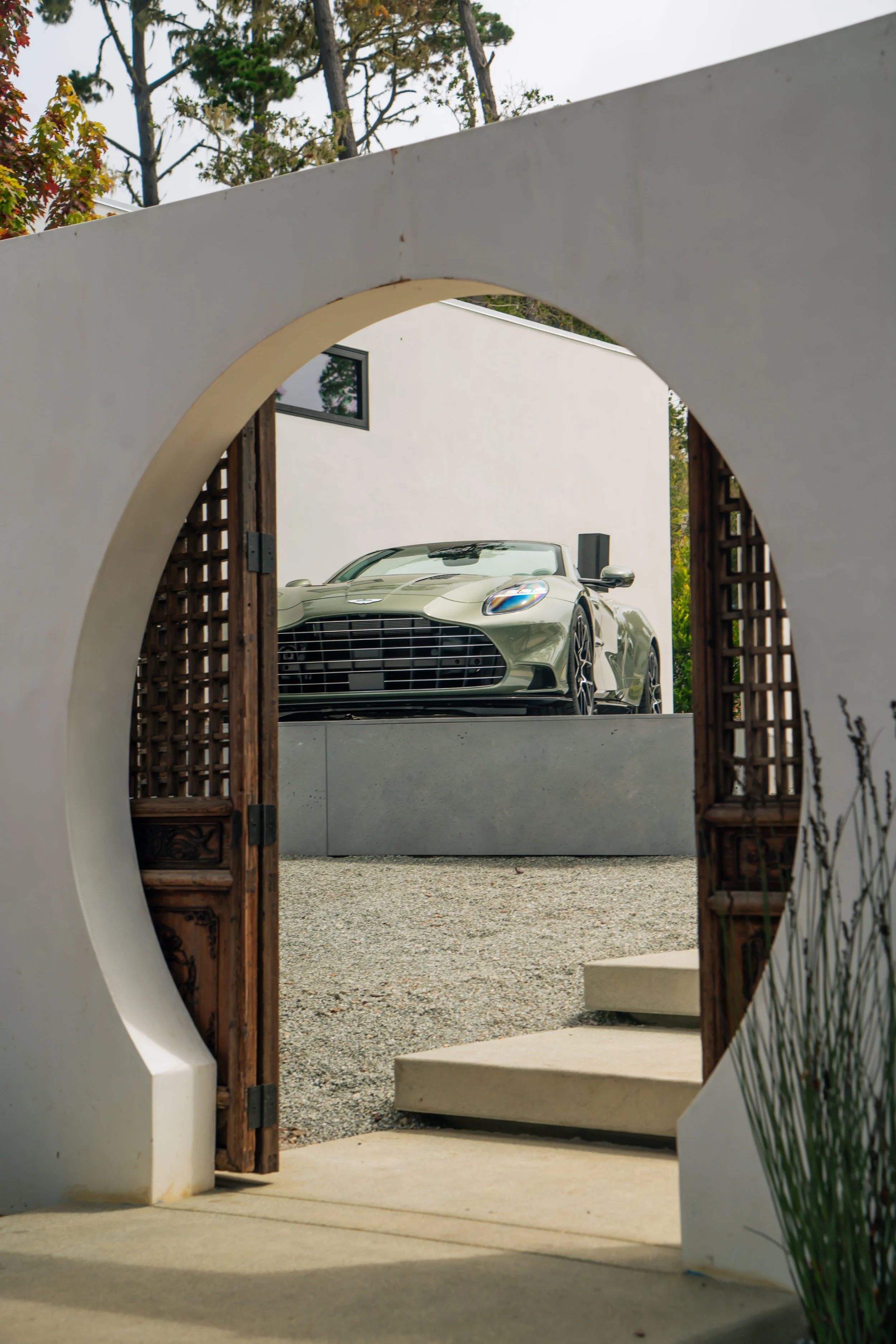 A luxury sports car parked outside a modern white building, viewed through a circular opening in a wall with a wooden accordion-style gate.