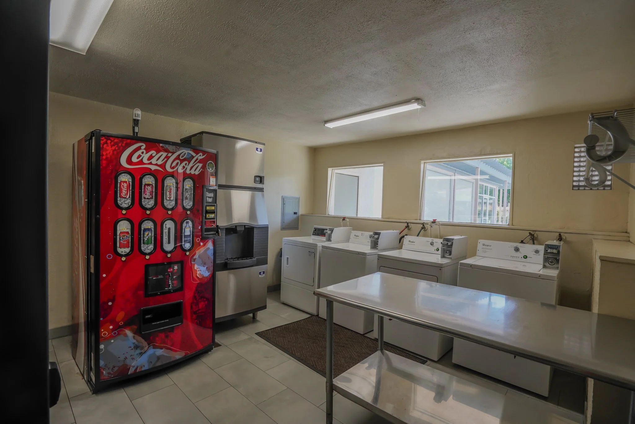 A small laundry room with a Coca-Cola vending machine on the left, a soda and ice machine beside it, three washers, and one dryer lined up against the wall underneath three windows. There is a stainless steel table in the foreground.