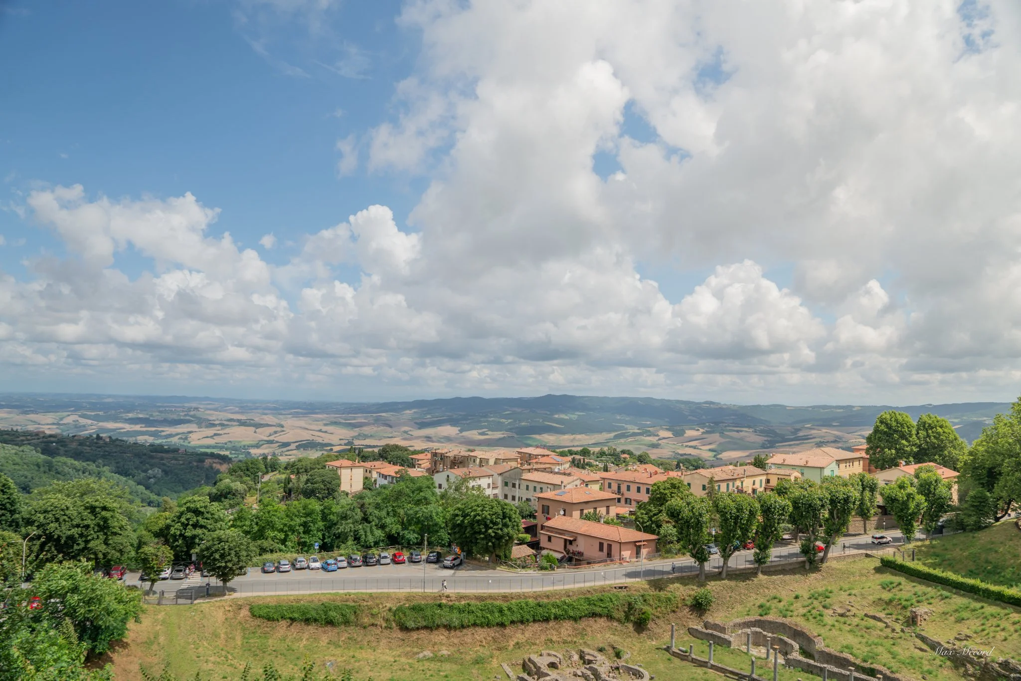 A scenic view of a small town with red-roofed buildings, surrounded by lush green trees and rolling hills, under a partly cloudy sky.