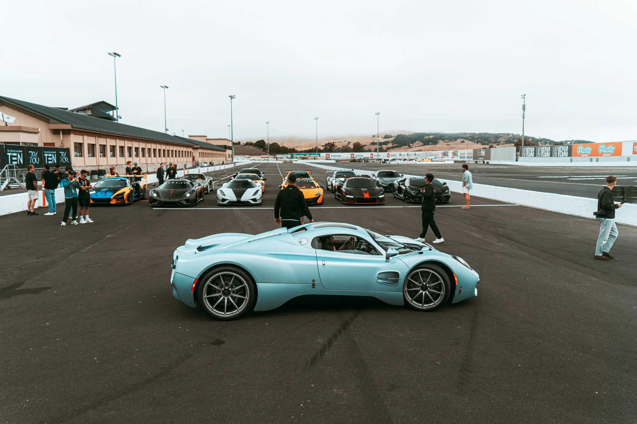 A row of luxury sports cars at a racetrack pit lane, with a light blue vintage car in the foreground, and a group of people, some taking photos and others observing, standing near the cars.