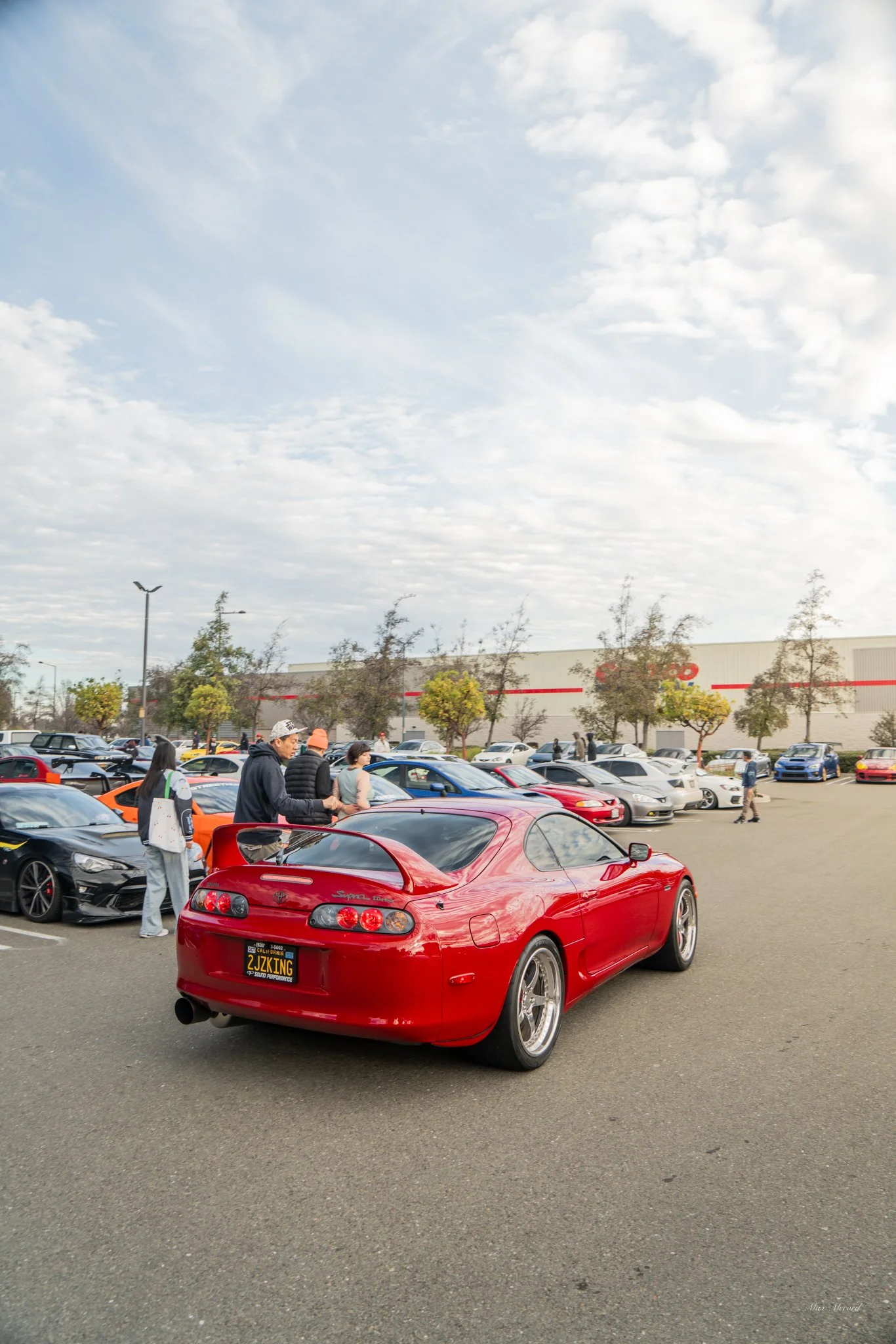 Red sports car in a parking lot with several people and other vehicles, against a partly cloudy sky.