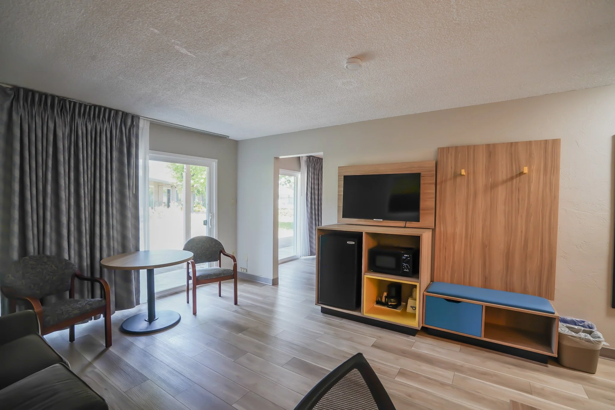 Living room with sliding glass door, wooden entertainment unit with TV, microwave, mini fridge, sitting area with chairs and small table, light flooring, white walls, gray curtains.