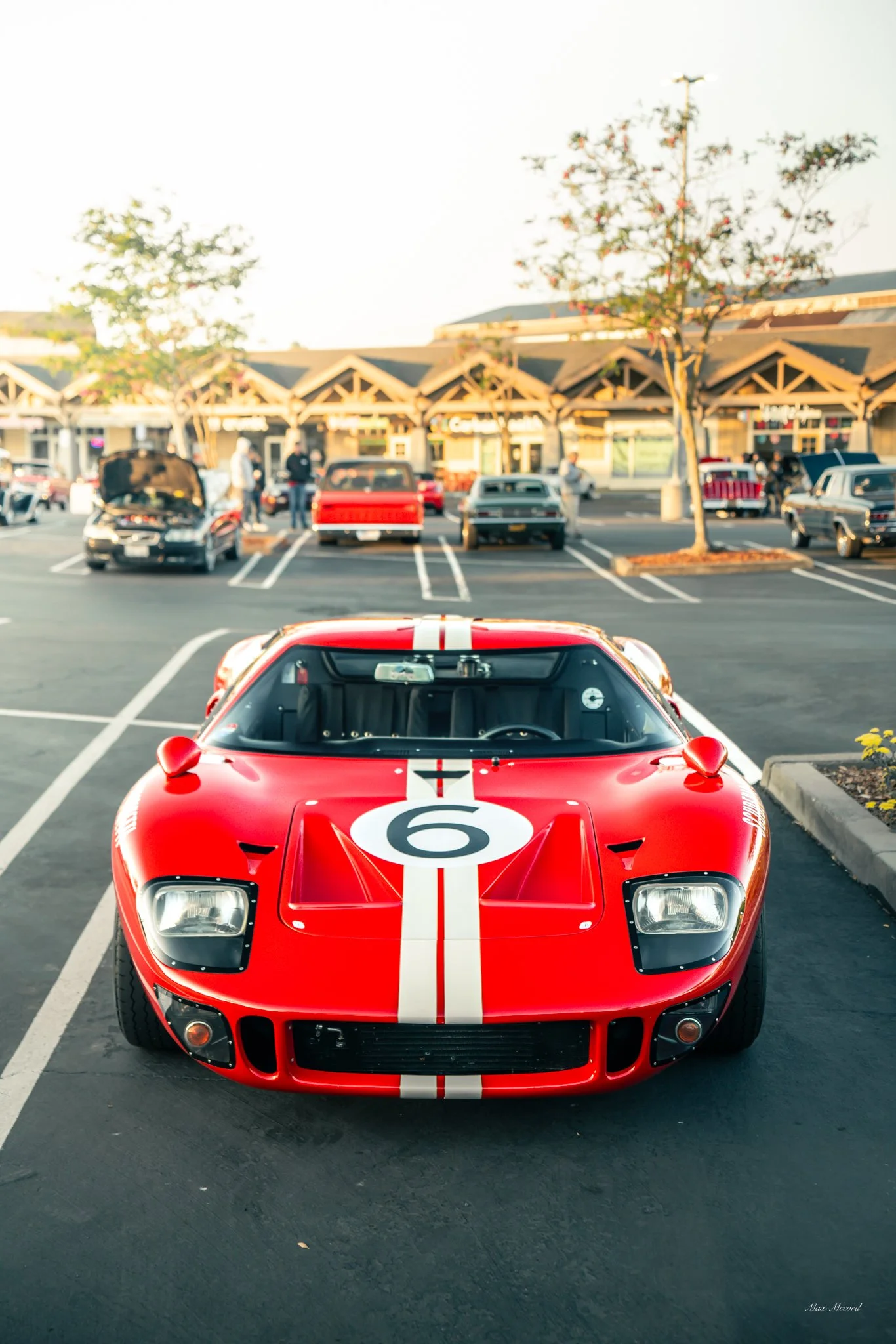 A red vintage race car with the number 6 on the hood, parked in a parking lot with a shopping center in the background.