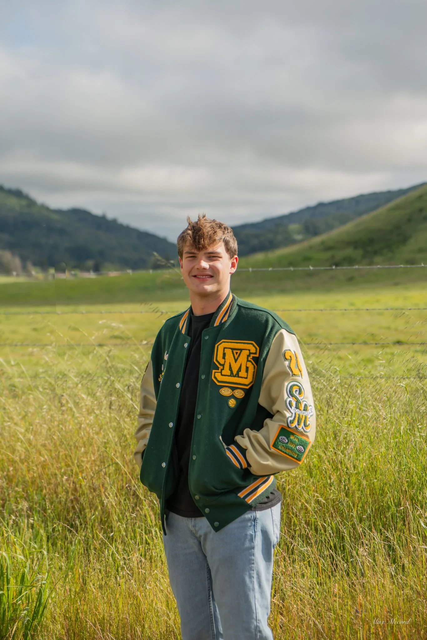 A young man standing in a grassy field outdoors, wearing a green and beige varsity jacket with patches, smiling at the camera, with rolling hills and a cloudy sky in the background.