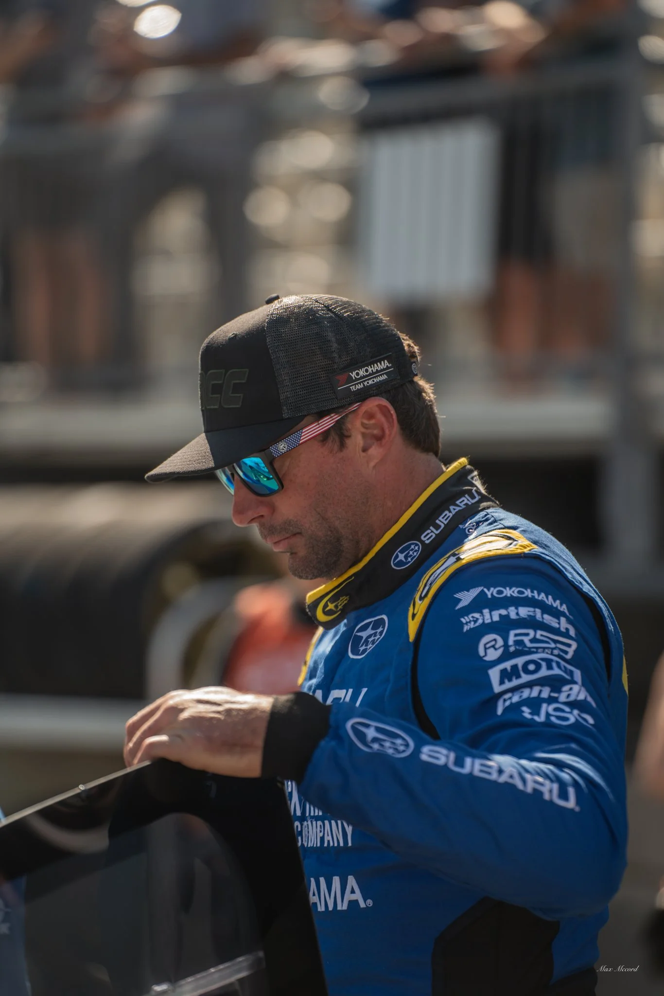 Race car driver in blue racing suit and sunglasses, adjusting something in his hand, outdoors with a blurred background.