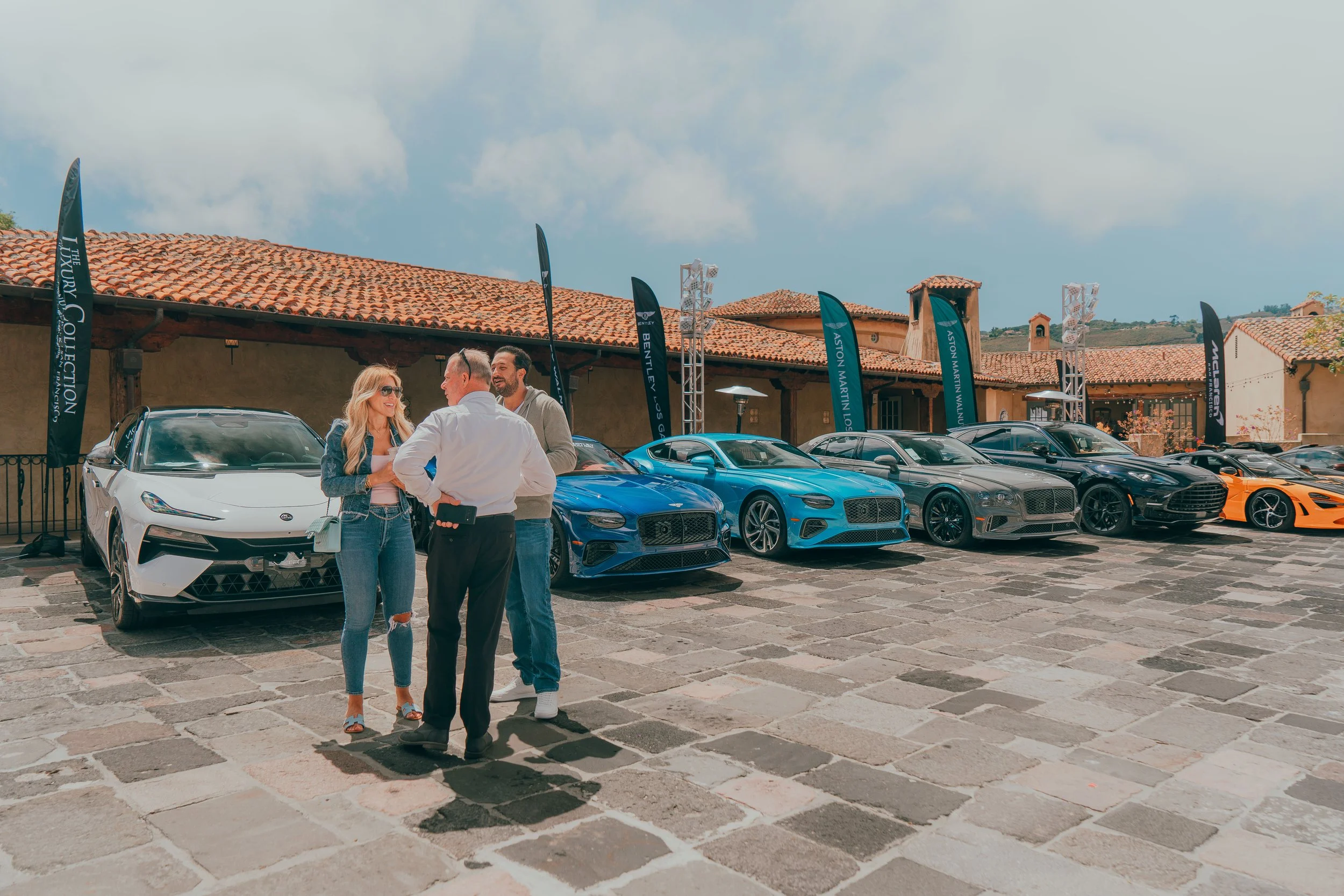 People talking in front of a row of luxury cars on display at an outdoor event with flags and a building with a tiled roof in the background.