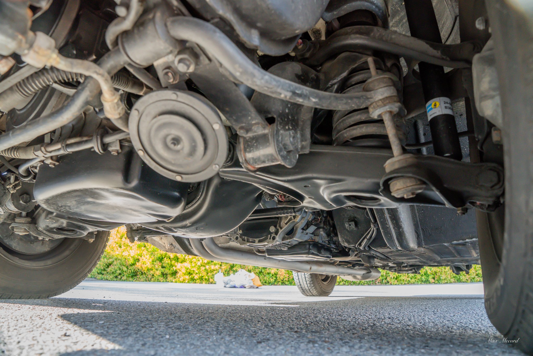 Underneath view of a car showing suspension components, shocks, and exhaust system on a paved surface with some greenery in the background.