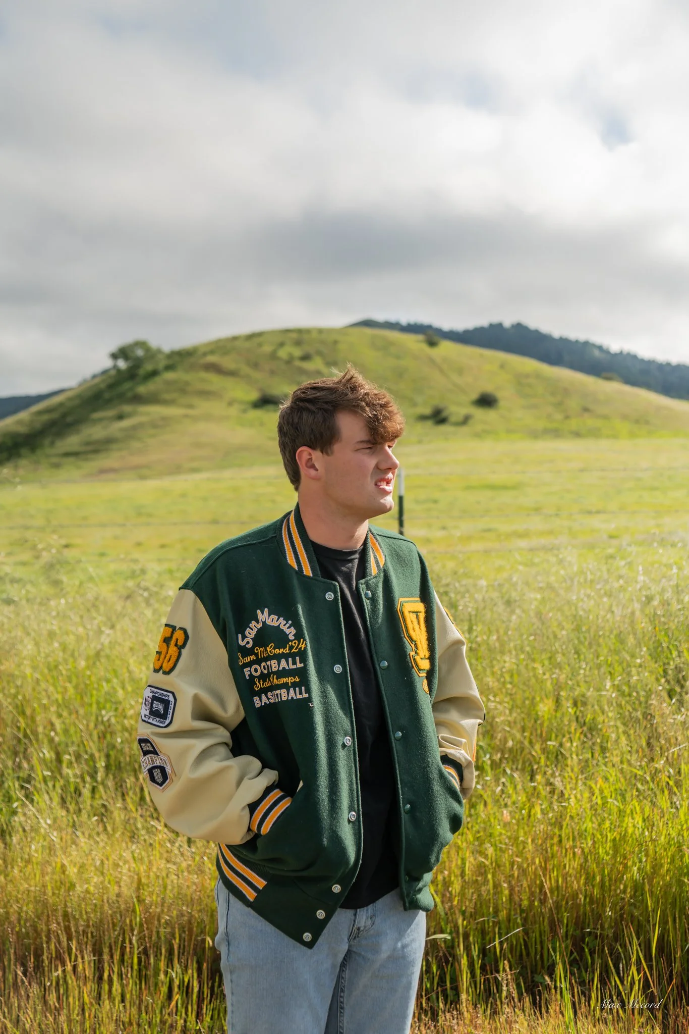 A young man with brown hair standing in a grassy field with a hill in the background, wearing a green and beige varsity jacket and looking to the right.