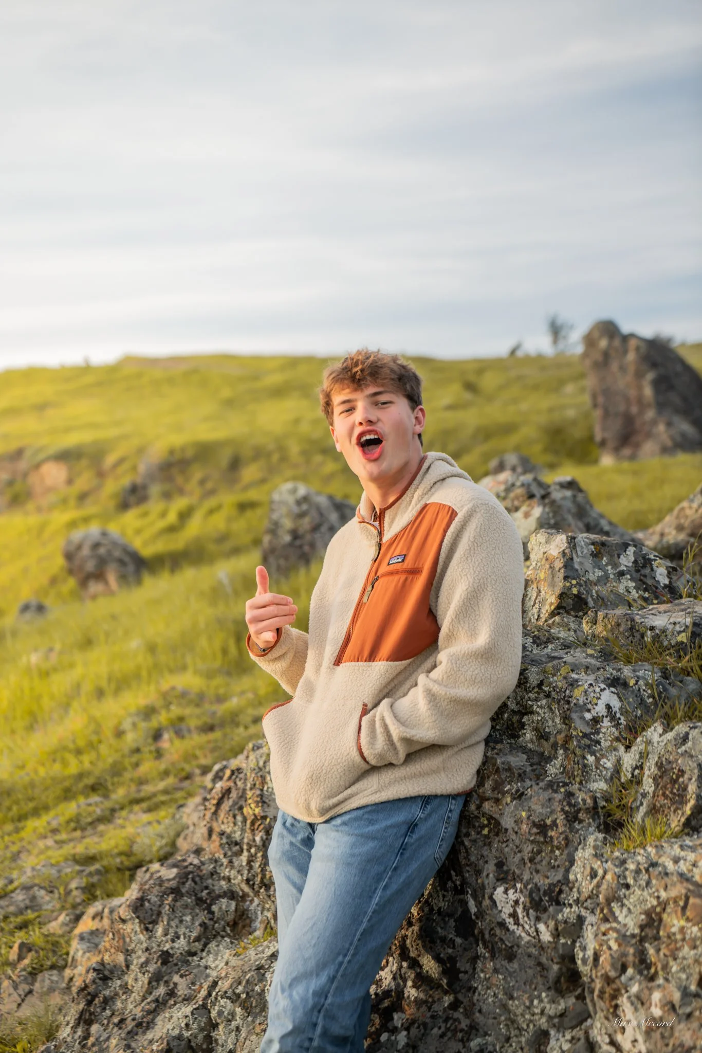 Young man with curly hair wearing a beige and orange Patagonia fleece jacket and blue jeans, leaning against rocks in a grassy, hilly landscape with a partly cloudy sky.