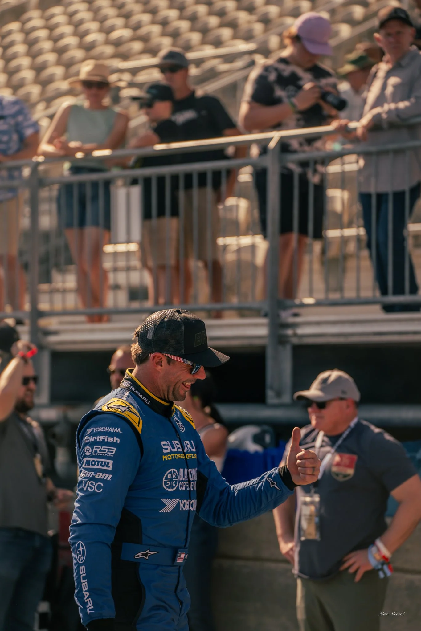 A race car driver in a blue Subaru racing suit with logos, giving a thumbs-up. He is smiling and wearing sunglasses and a cap. People in the background and spectators on a balcony watch, some taking photos.