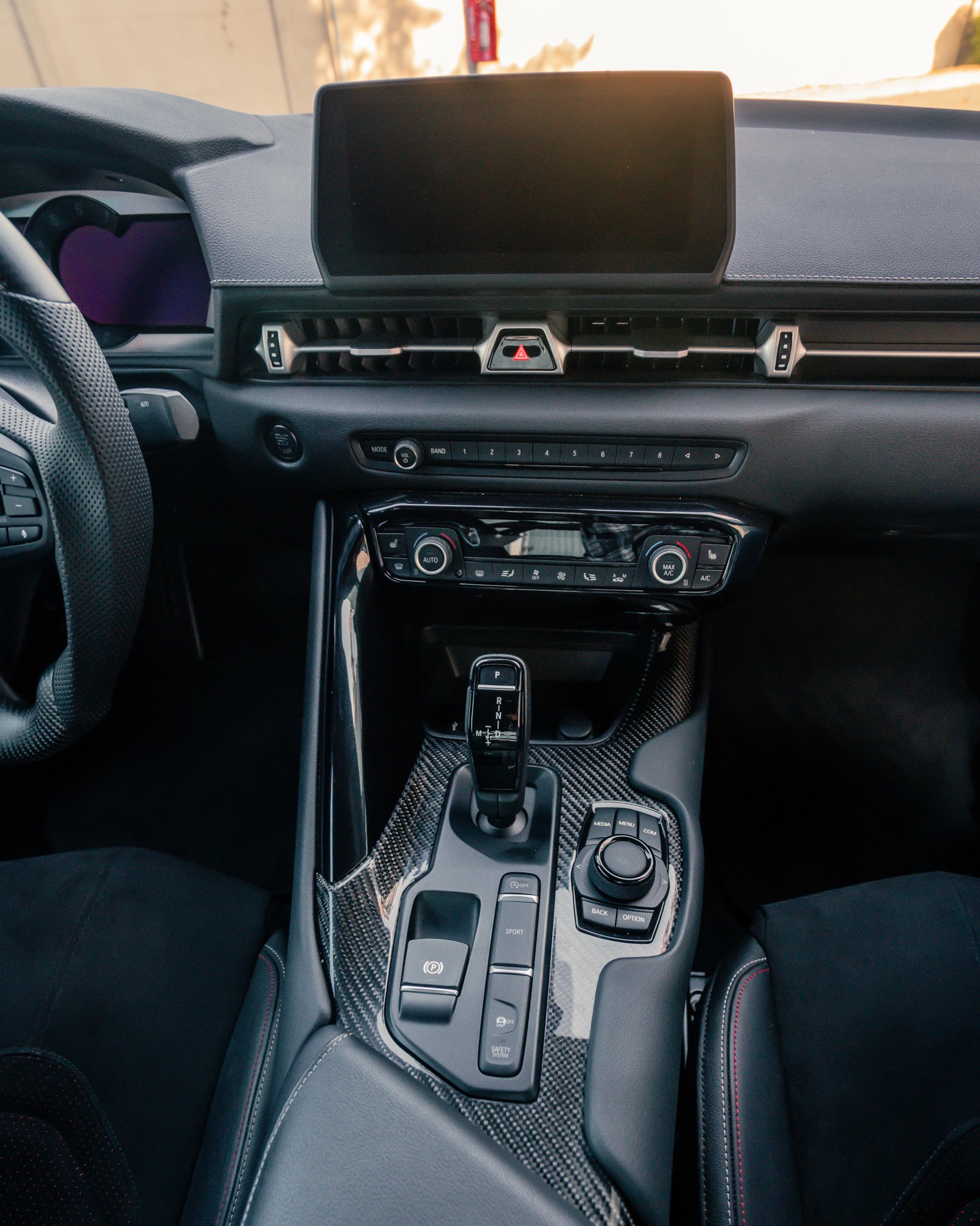 The interior of a modern car, featuring a dashboard with a large central display, climate control buttons, a gear shift, and control knobs on a carbon fiber console.
