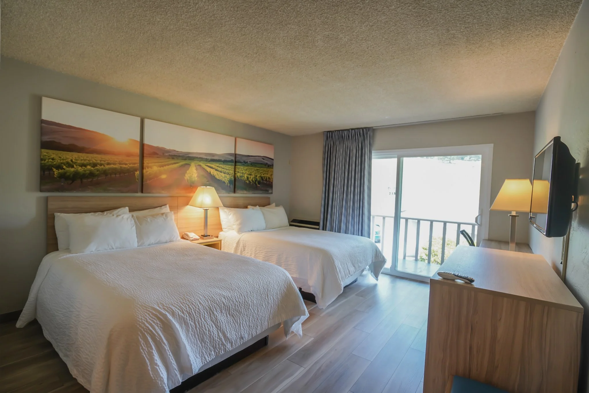 A hotel room with two double beds with white bedding, a wall art of a vineyard at sunset above the beds, a nightstand with a lamp between the beds, a window with blue curtains leading to a balcony, and a dresser with a TV and a remote.