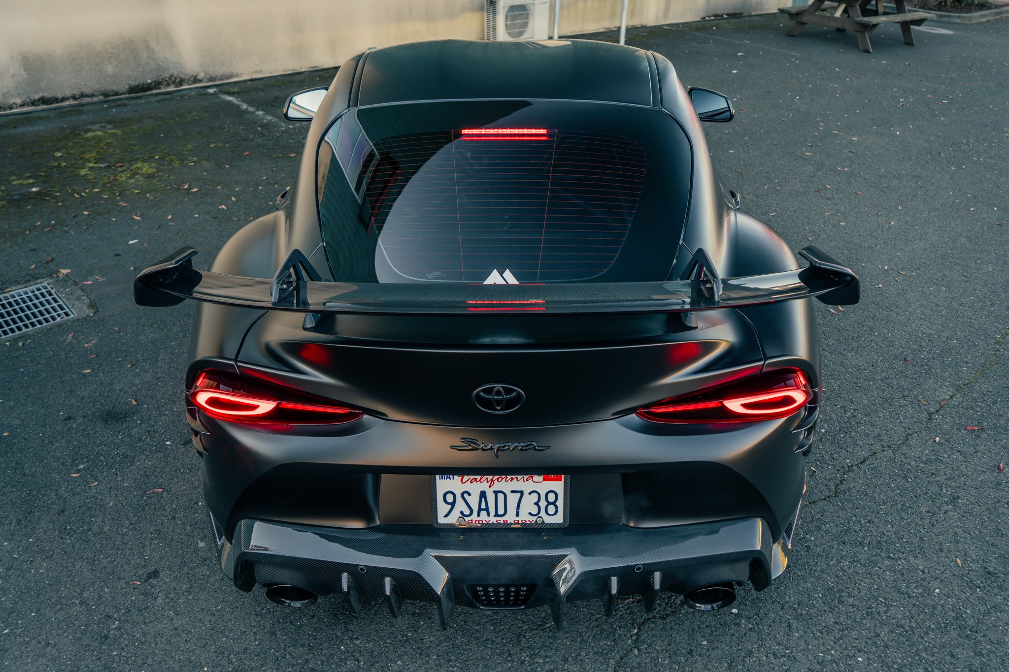 Top view of a black Toyota Supra sports car parked on asphalt, featuring a large rear wing, red tail lights, and dual exhaust pipes.