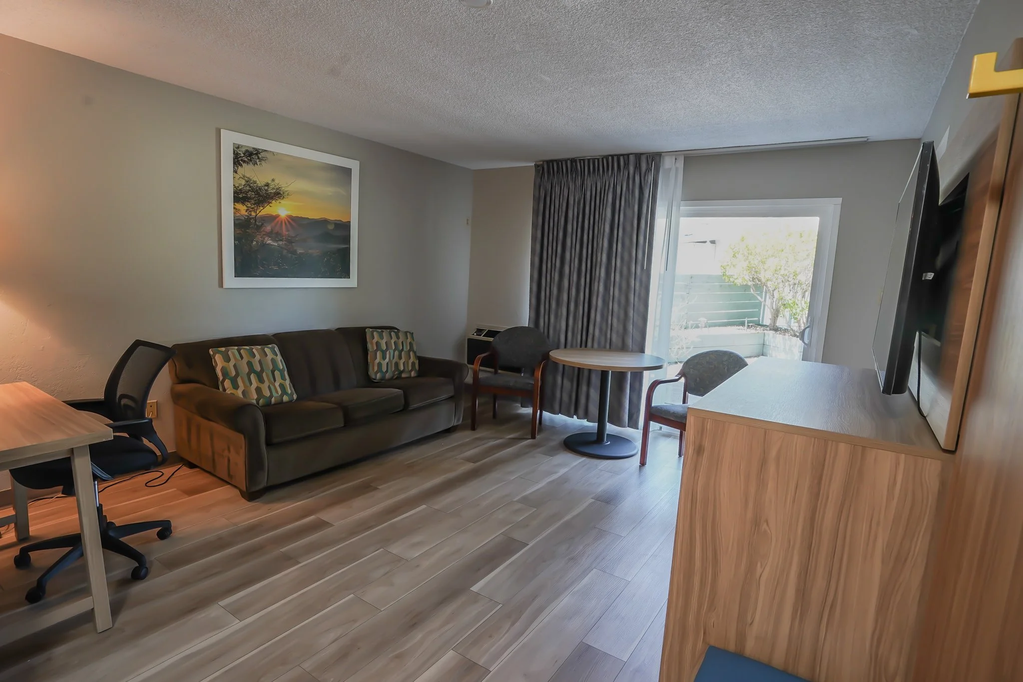 Living room with a dark sofa, patterned pillows, a small round table, a desk with an office chair, a flat-screen TV on a wooden stand, a sliding glass door with curtains, and artwork of a sunset on the wall.