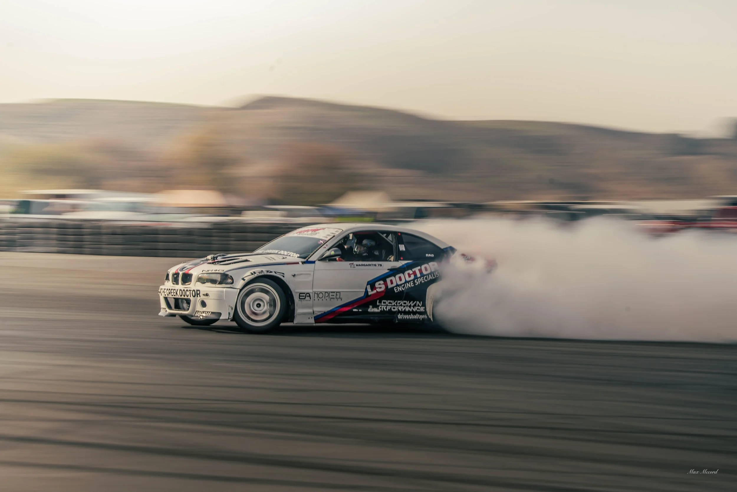 A silver race car drifting on a race track, creating smoke from the tires, with a blurred background of hills and other vehicles.