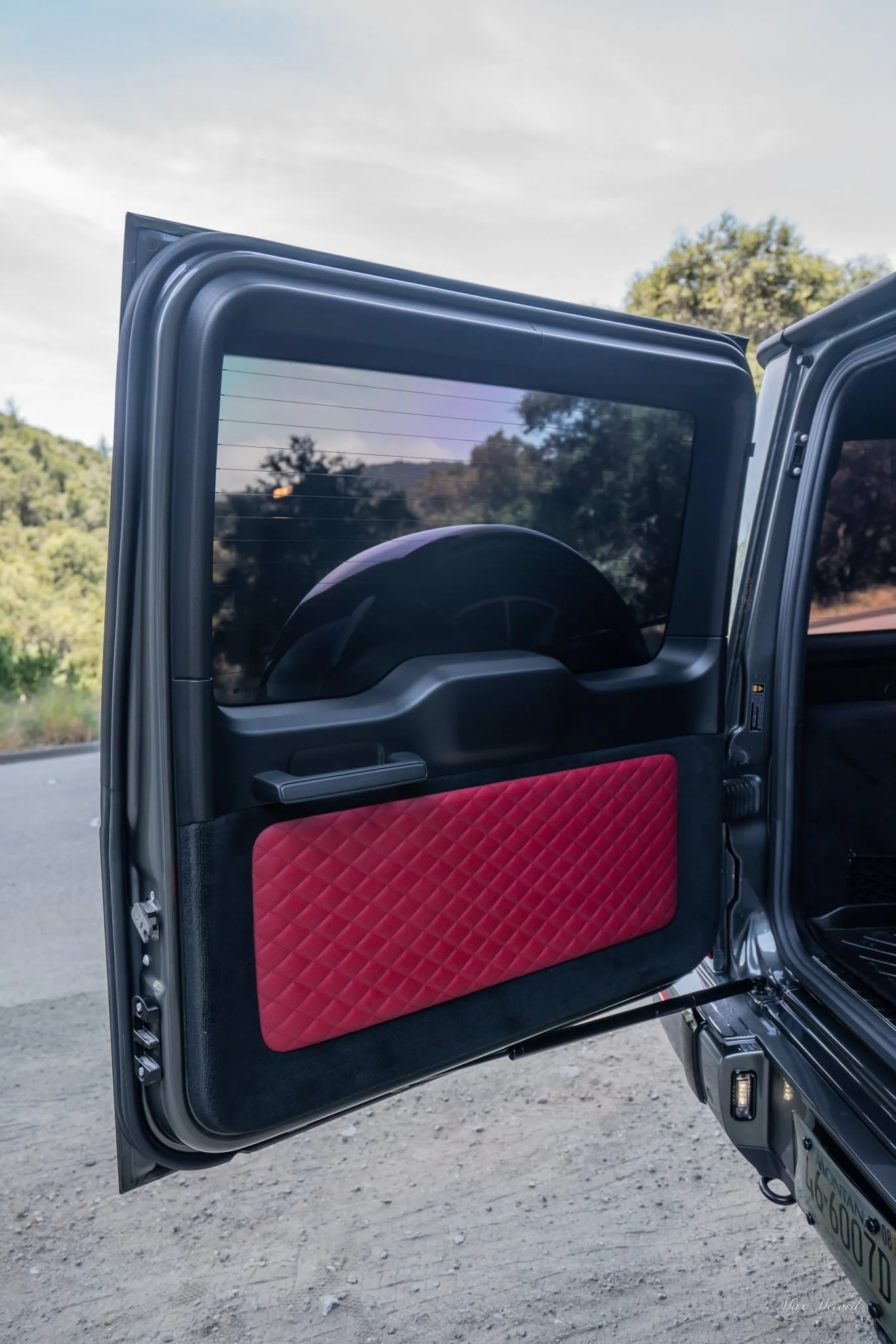 The open rear door of a black SUV with red quilted interior panel, showing a mountain bike inside, on a dirt road with lush trees and mountains in the background.