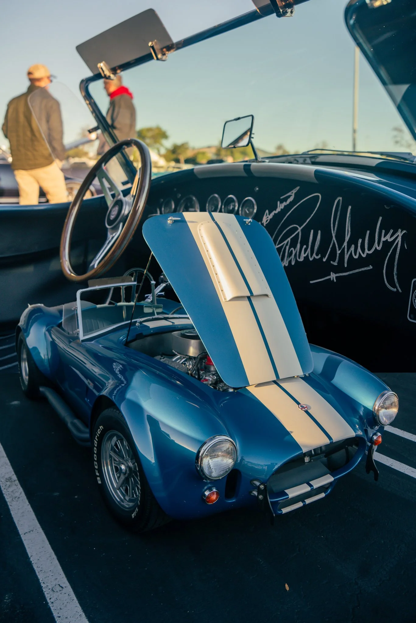 A miniature blue and white vintage race car with its hood open on display at a car show, with two people and a classic car in the background.