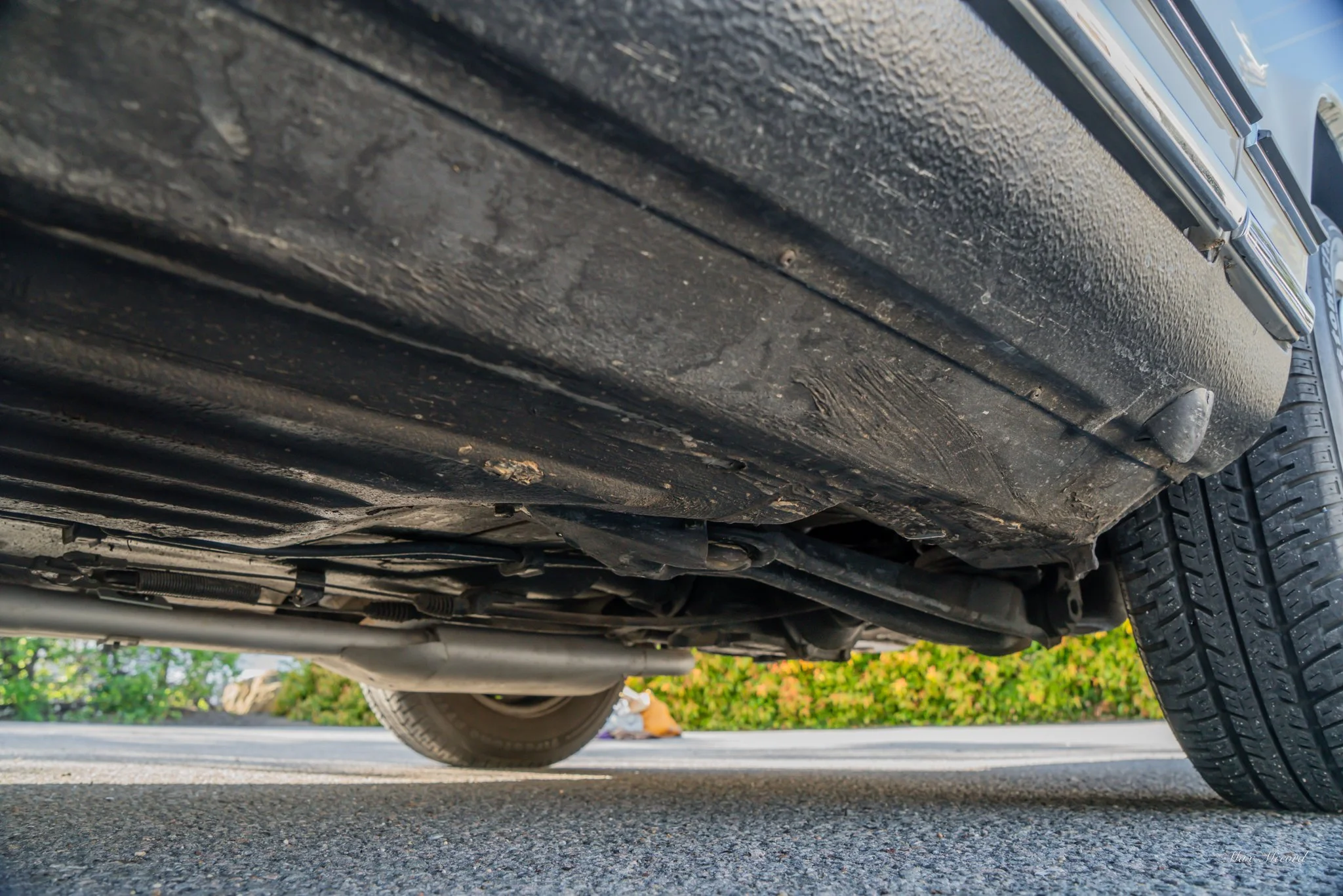 Low-angle view of the underside of a vehicle parked on the street, showing the undercarriage, tire, and pavement.
