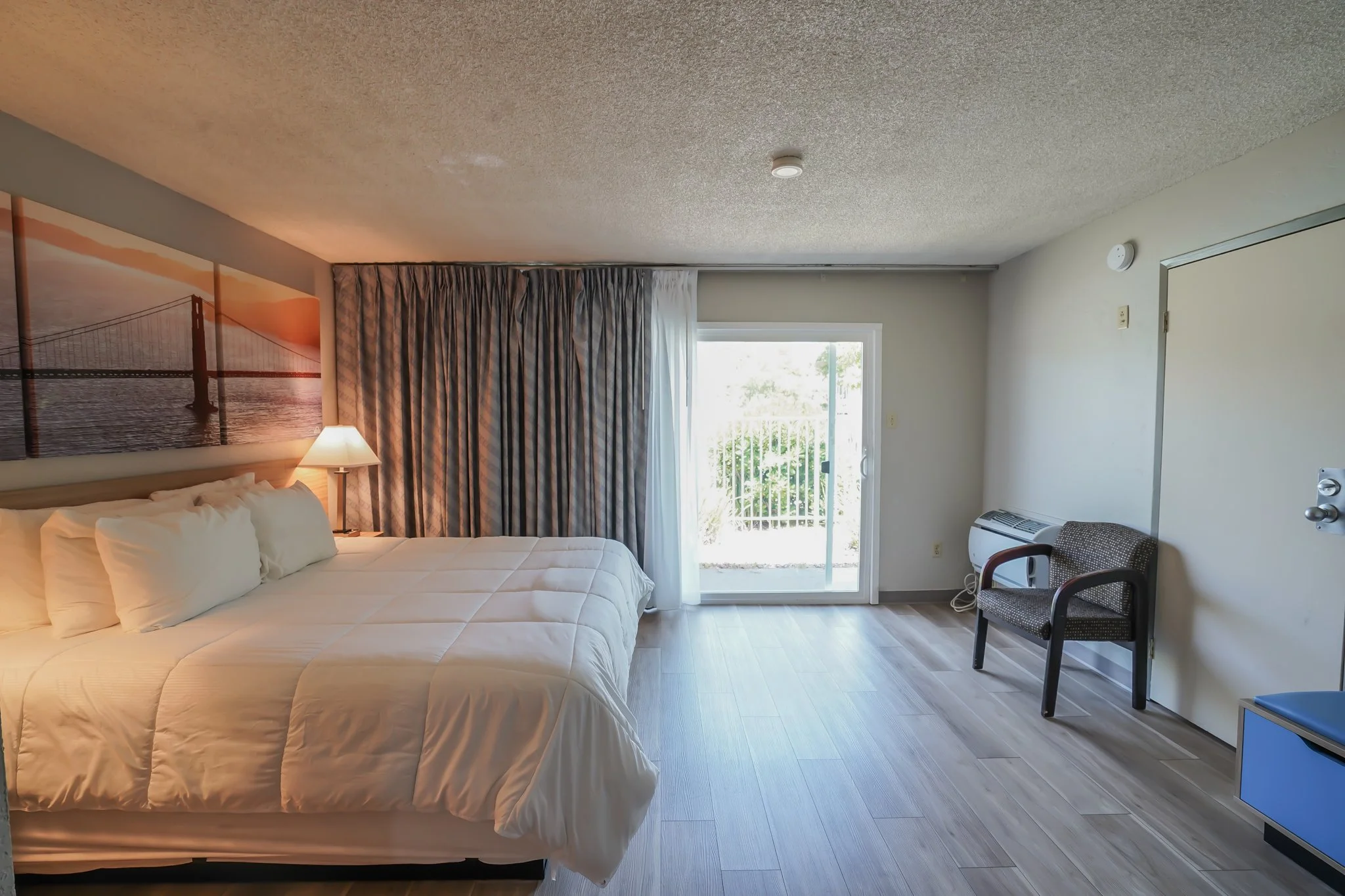 A hotel room with a king-sized bed, a nightstand with a lamp, artwork of a bridge at sunset, curtains partially open to a balcony door, a chair, and an air conditioning unit.