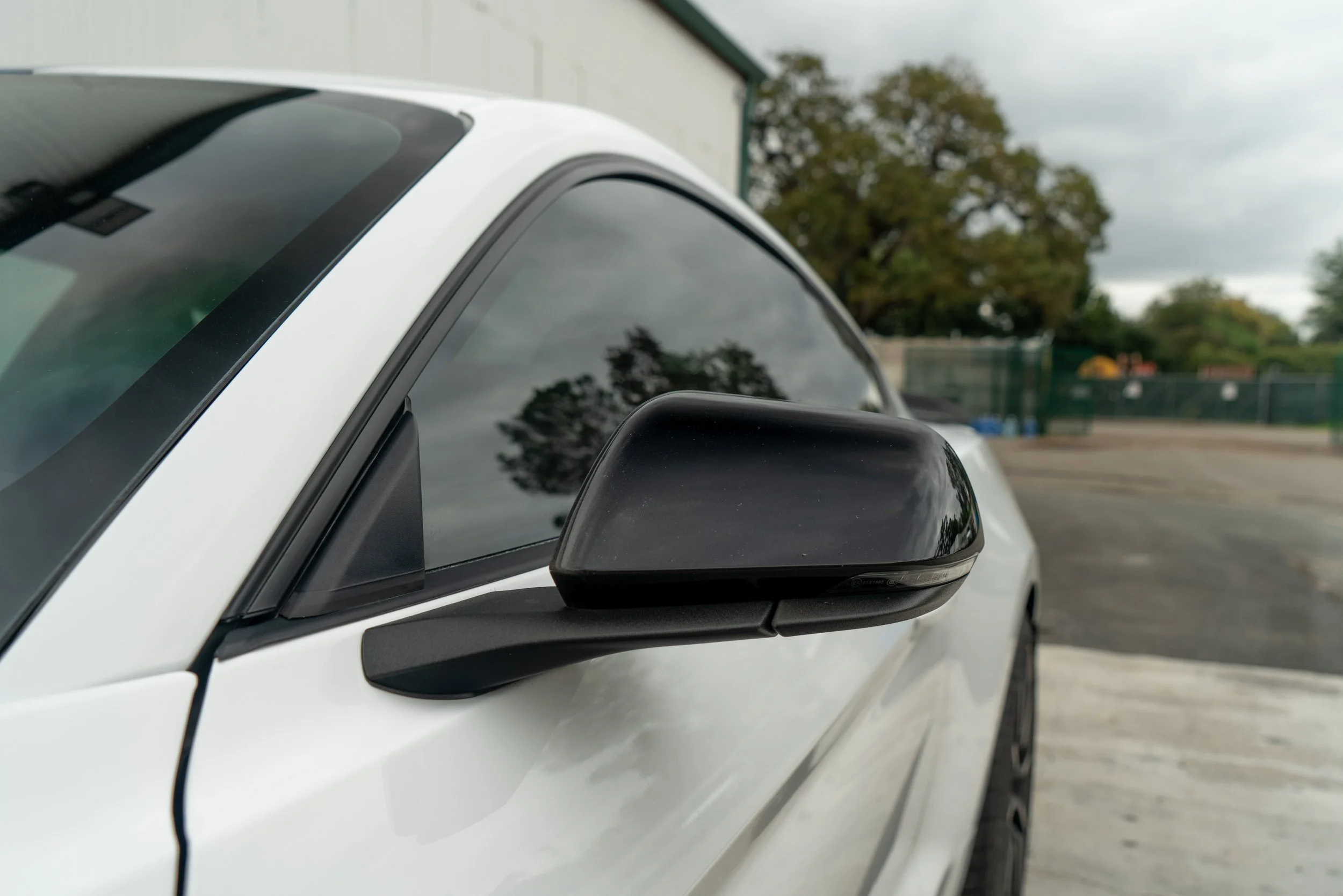Close-up of a white car's black side mirror and front passenger window, with trees and a parking lot in the background under cloudy skies.