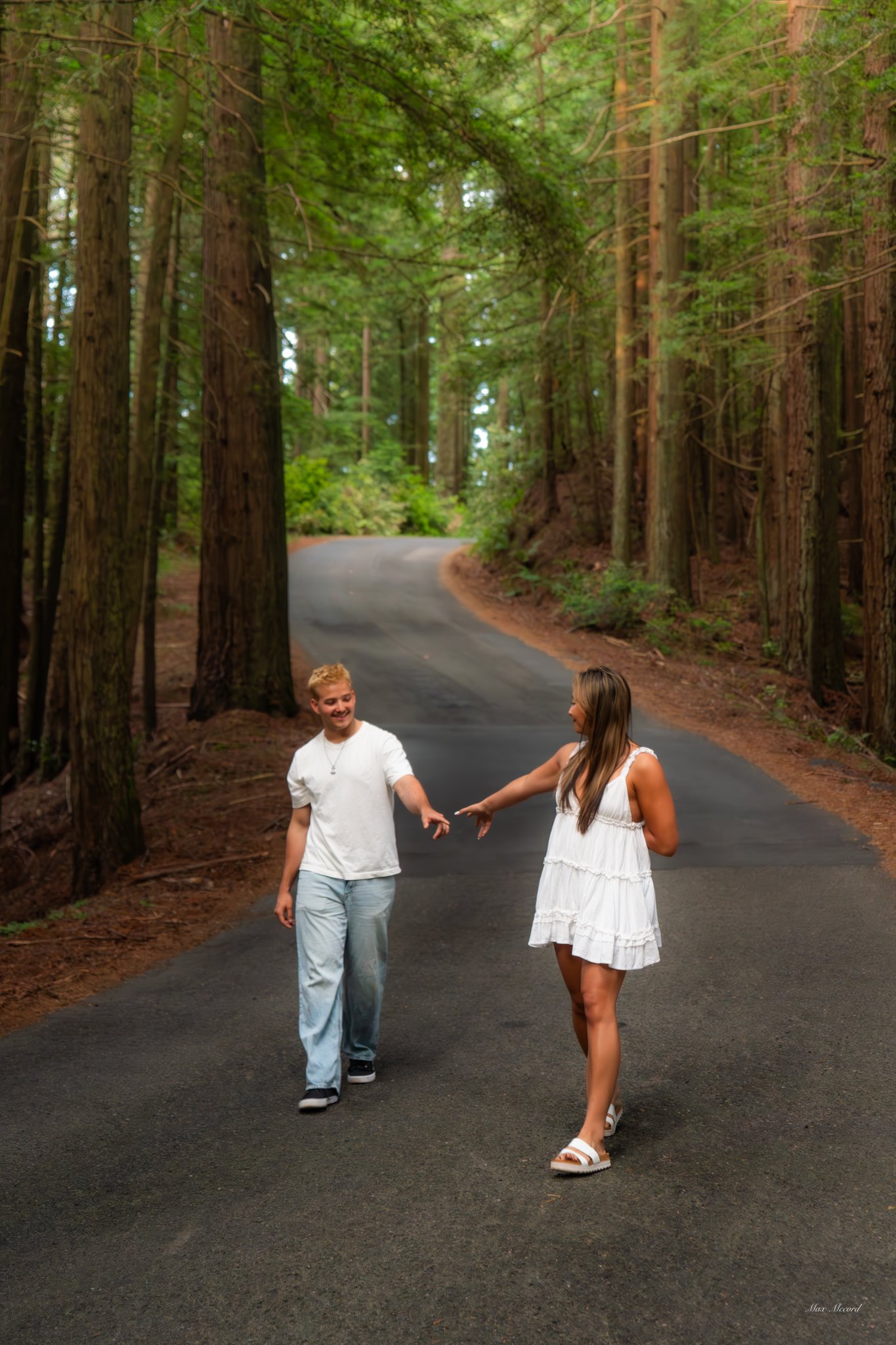 A young man and woman are walking and playing on a winding paved road through a forest with tall trees and green foliage, smiling and reaching out to each other.