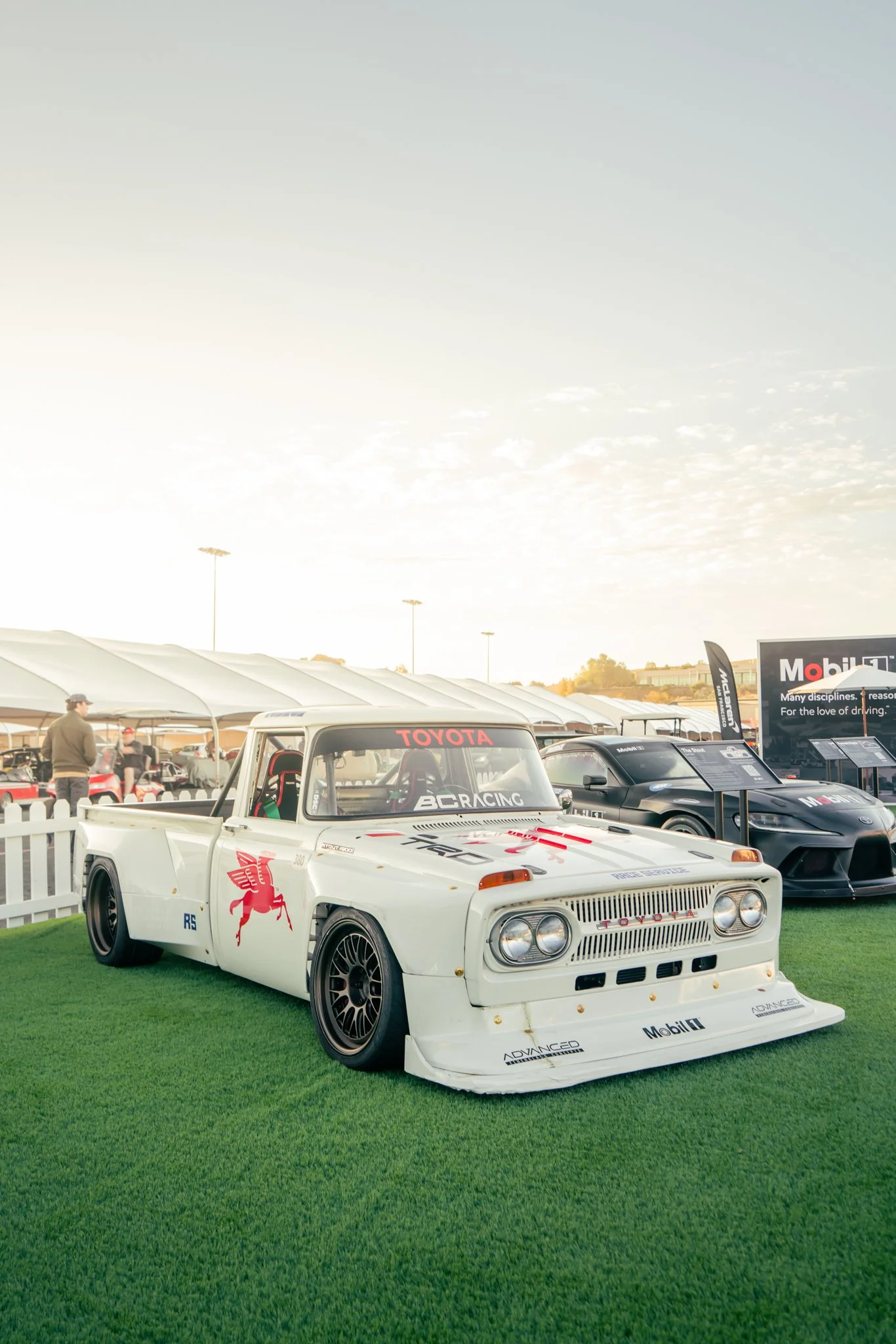 A vintage white Toyota race car with red and black decals parked on green grass at an outdoor car event, with tents, people, and modern cars in the background.