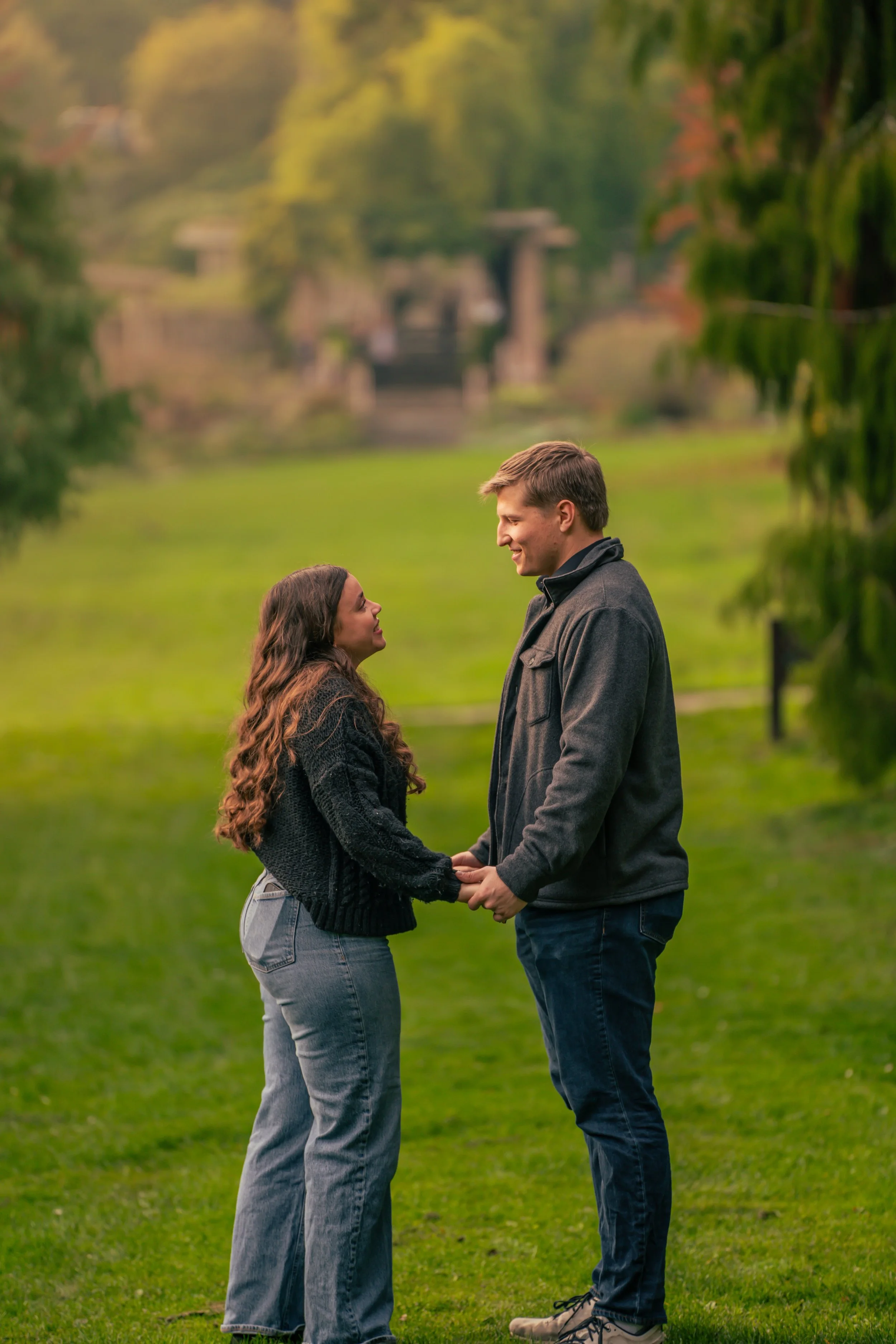 A couple holding hands and smiling at each other in a park with green grass and trees, with a blurred background of a river and structures.