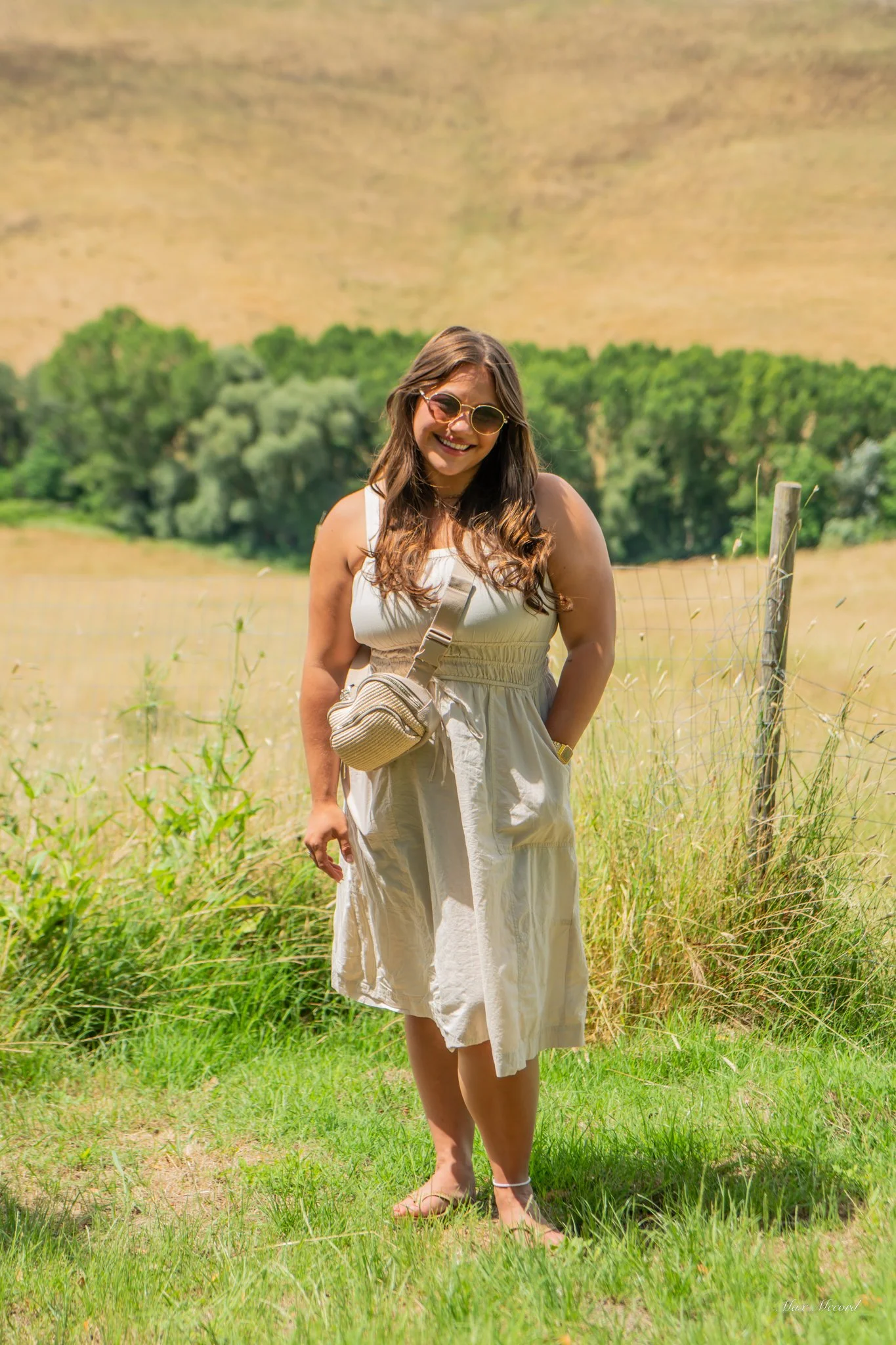 A woman with long wavy hair, sunglasses, and a white summer dress, smiling outdoors in a grassy field with trees and rolling hills in the background.